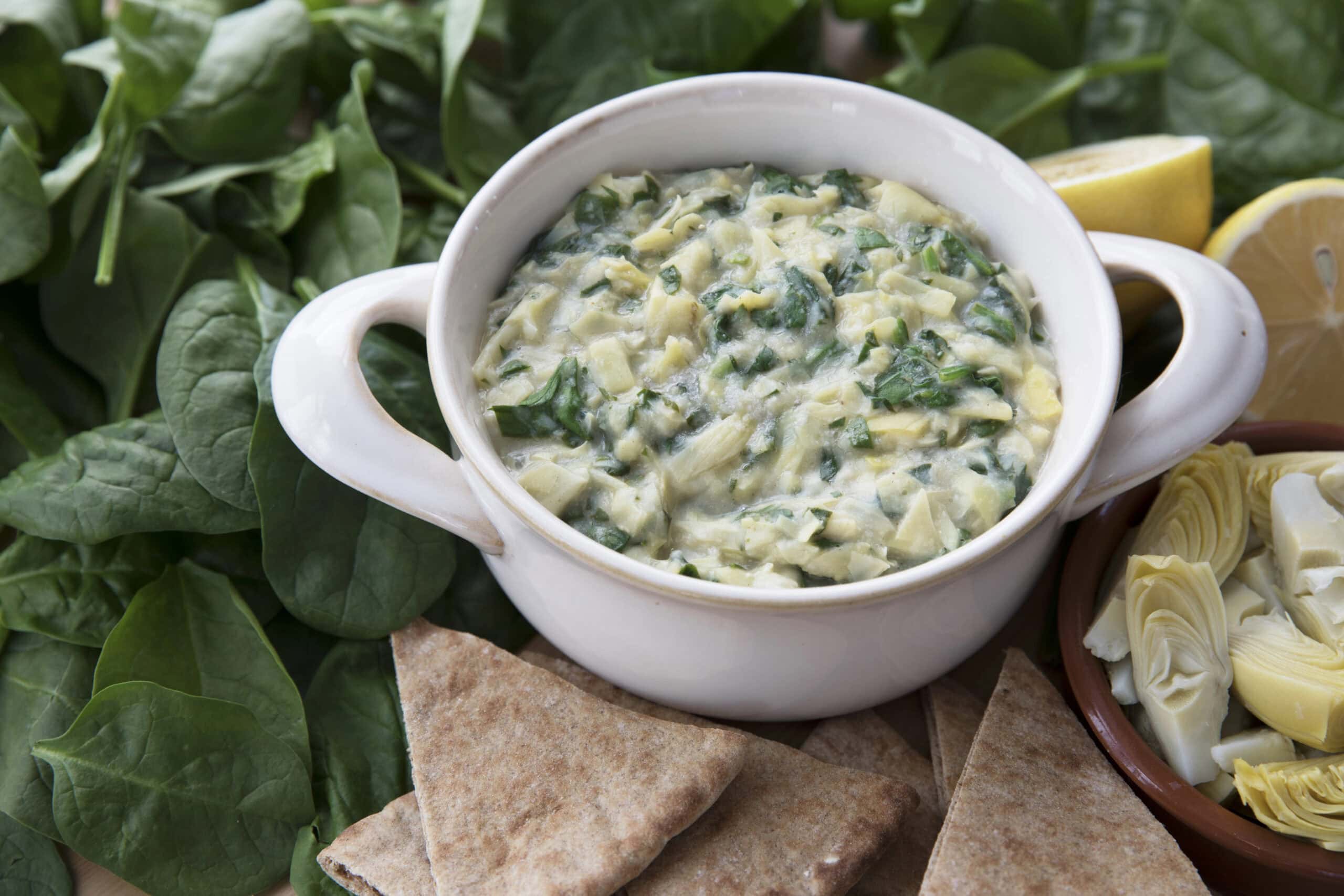 A close-up of spinach and artichoke dip in a white serving bowl surrounded by fresh spinach leaves, a bowl of artichoke hearts, and whole wheat pita bread triangles.