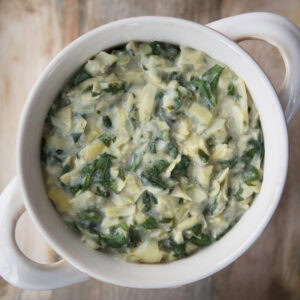 A top-down view of a white bowl filled with spinach and artichoke dip on a rustic wooden surface.