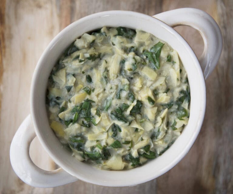 A top-down view of a white bowl filled with spinach and artichoke dip on a rustic wooden surface.