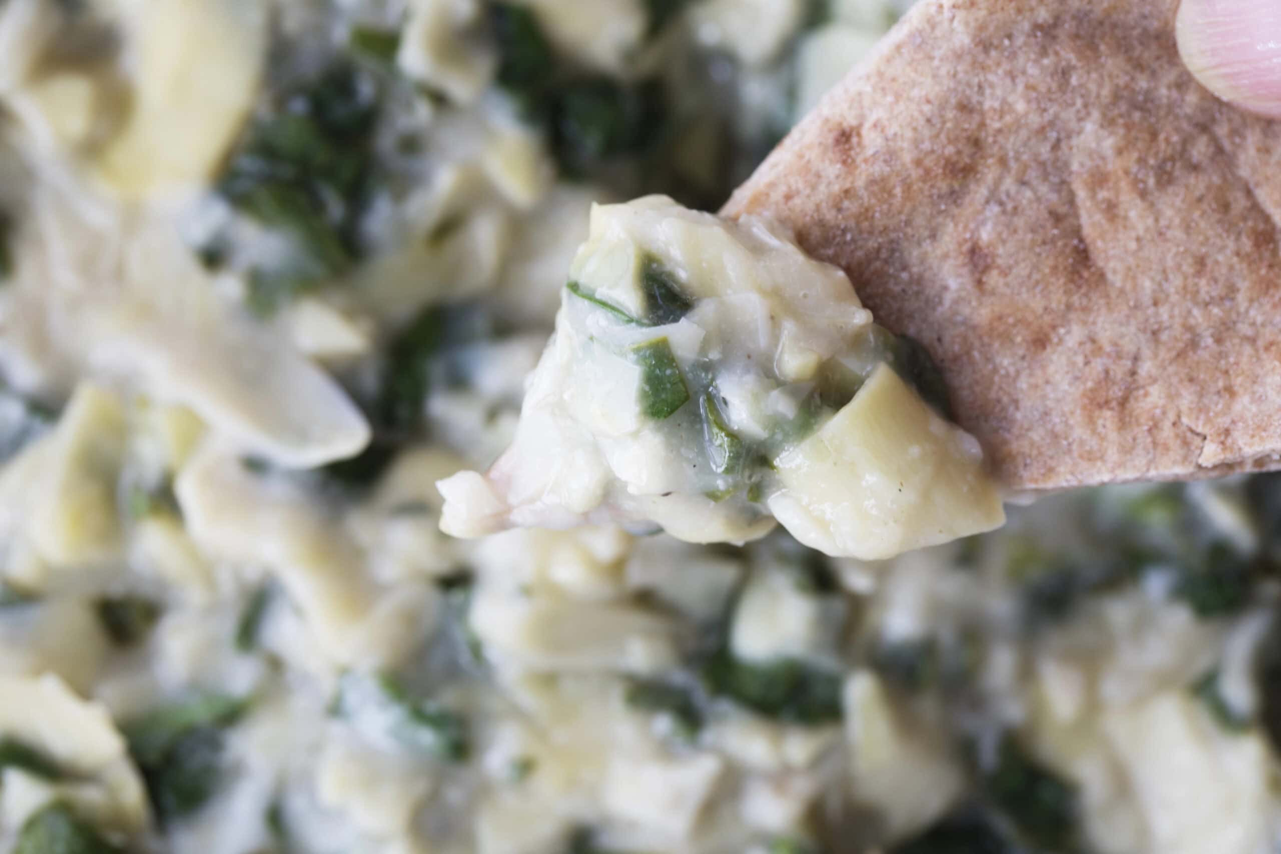 A close-up of a piece of pita bread dipped into a creamy spinach and artichoke dip, highlighting the textures of the ingredients.