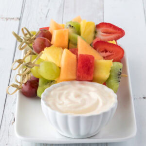 A white plate featuring colorful fruit skewers with pineapple, watermelon, strawberries, grapes, and kiwi alongside a bowl of Greek yogurt dip, set on a rustic white table.
