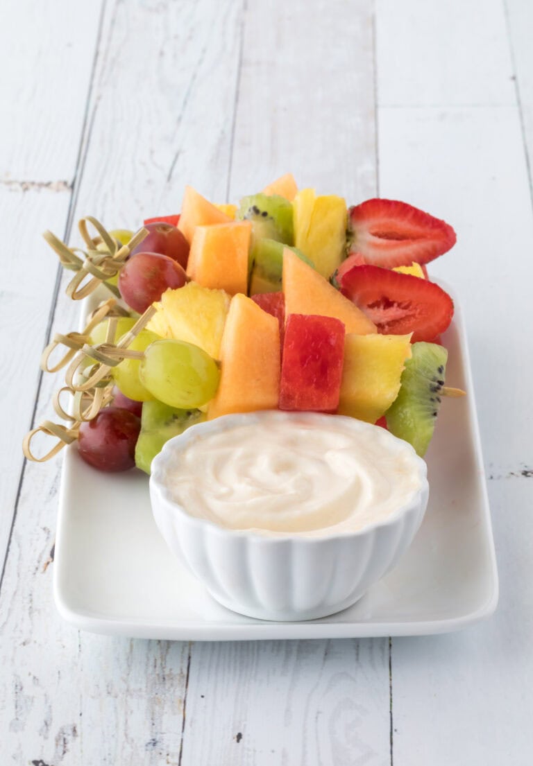 A white plate featuring colorful fruit skewers with pineapple, watermelon, strawberries, grapes, and kiwi alongside a bowl of Greek yogurt dip, set on a rustic white table.