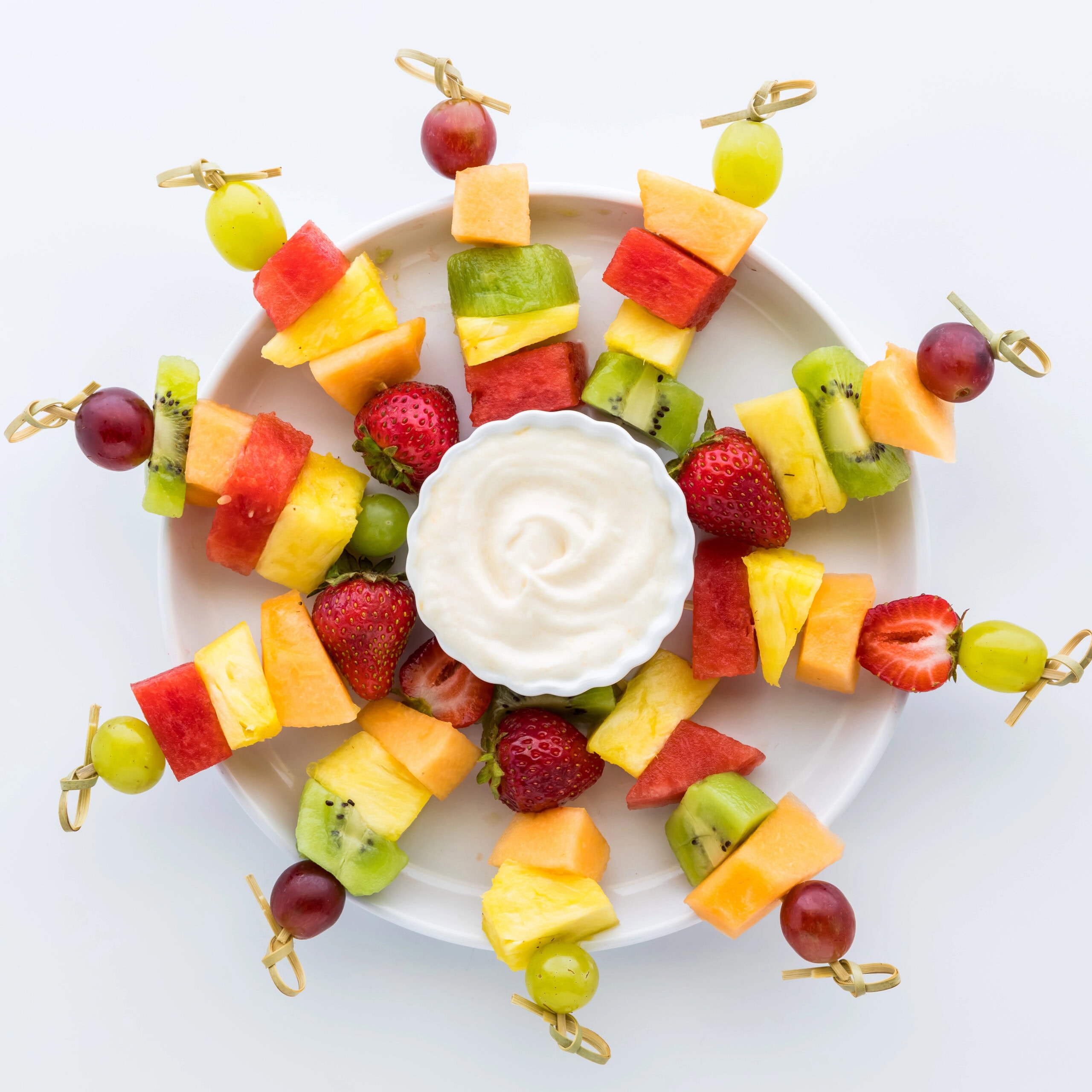 A round fruit platter arranged with skewers of watermelon, strawberries, pineapple, grapes, and kiwi encircling a bowl of yogurt dip, styled on a white background.