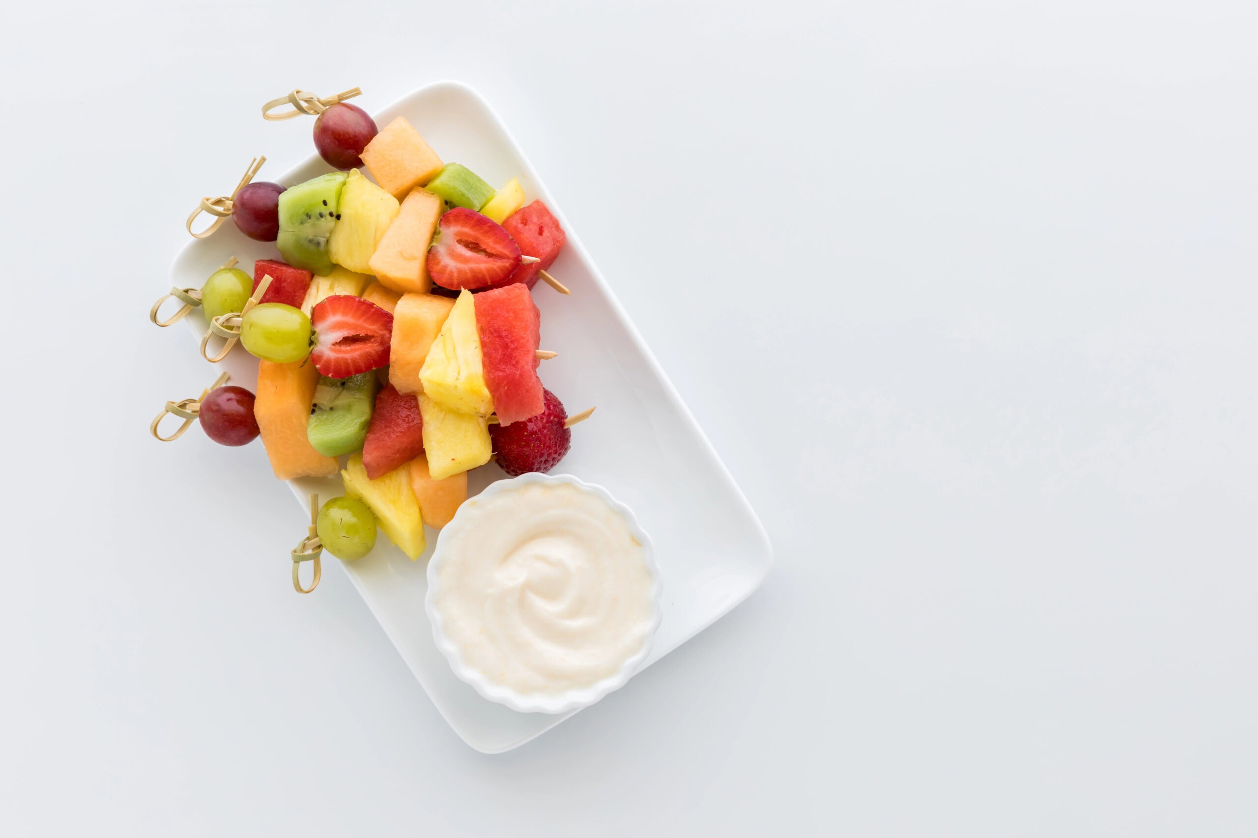 A minimalist presentation of fruit skewers with vibrant colors including pineapple, watermelon, strawberries, grapes, and kiwi paired with a bowl of yogurt dip on a white rectangular plate.