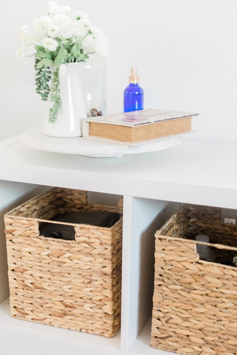 A stylish storage unit featuring white cubby shelves with woven wicker baskets for organizing small spaces. The top of the unit is decorated with a white ceramic vase holding faux flowers, a wooden book, and a small blue spray bottle.
