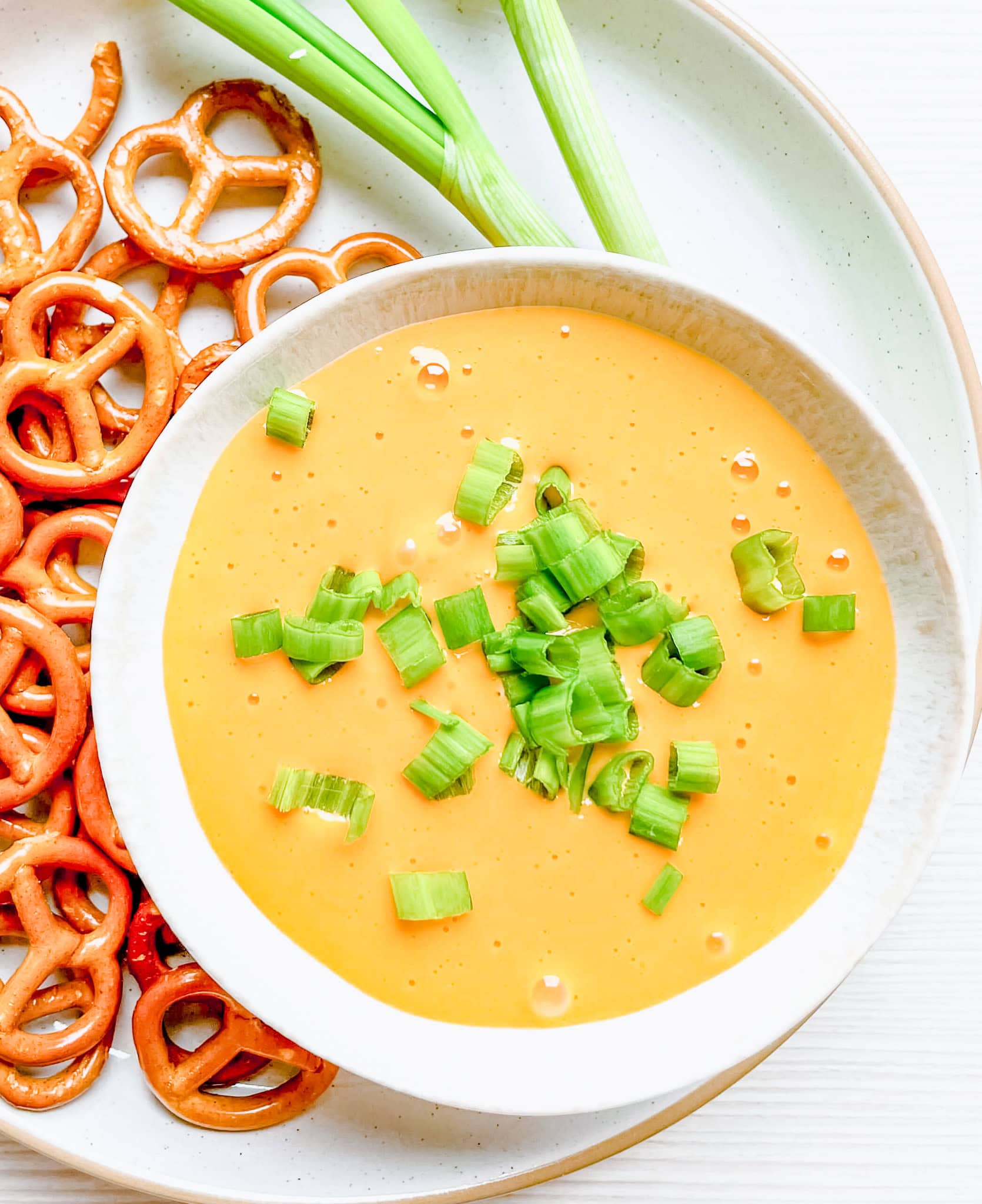 A bowl of hot beer and cheese dip garnished with freshly chopped green onions, surrounded by golden pretzels and fresh green onions on a light-colored plate. The dip is creamy and smooth, with a vibrant orange color.