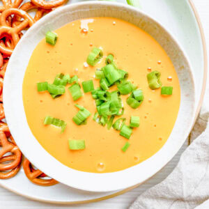 A wider shot of the hot beer and cheese dip, placed on a light-colored plate with pretzels and fresh green onion stalks. A beige linen napkin is partially visible on the side.