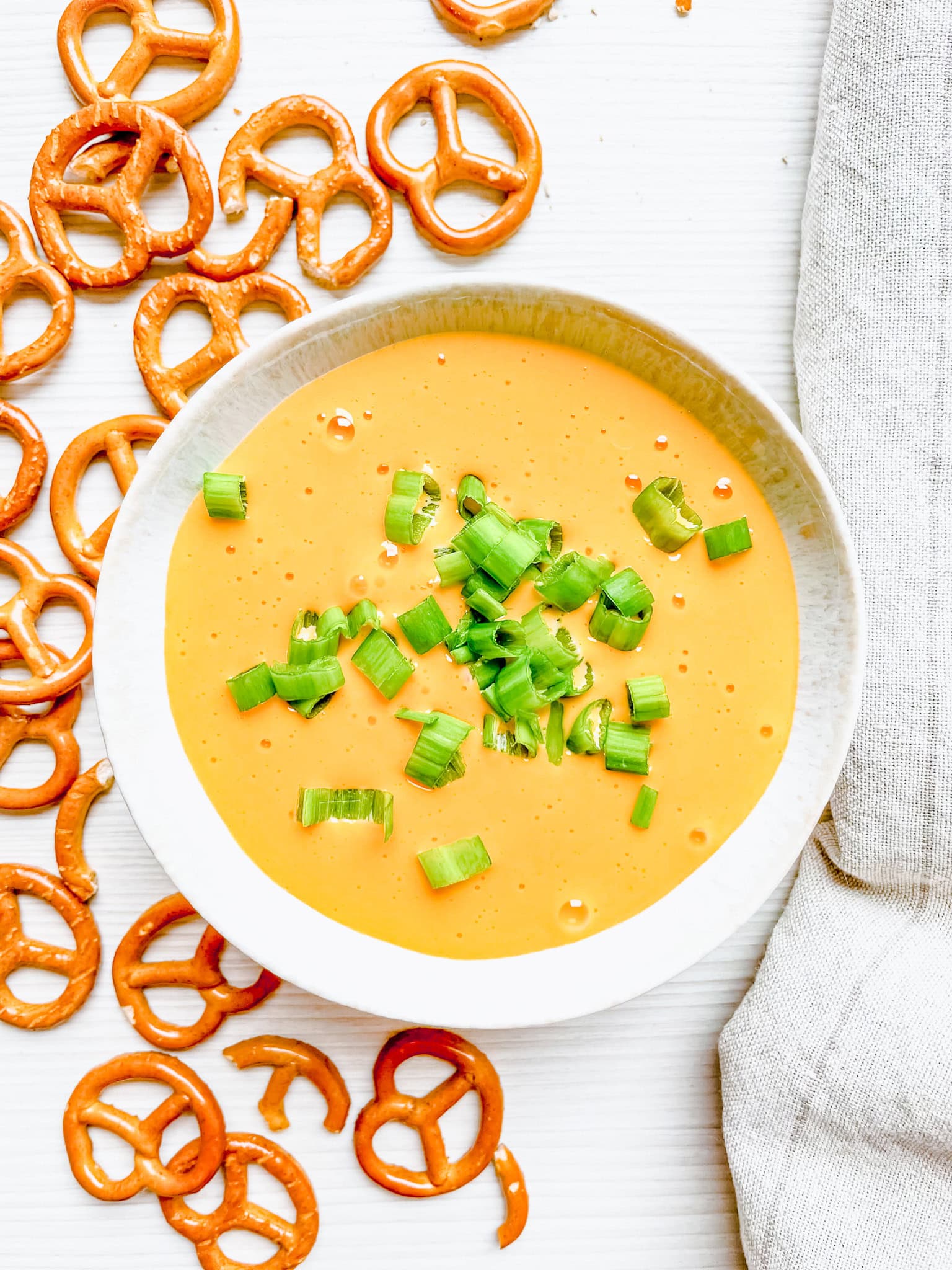 Overhead view of the hot beer and cheese dip in a white bowl, garnished with chopped green onions. Surrounding the bowl are scattered pretzels and a linen napkin on a white surface.