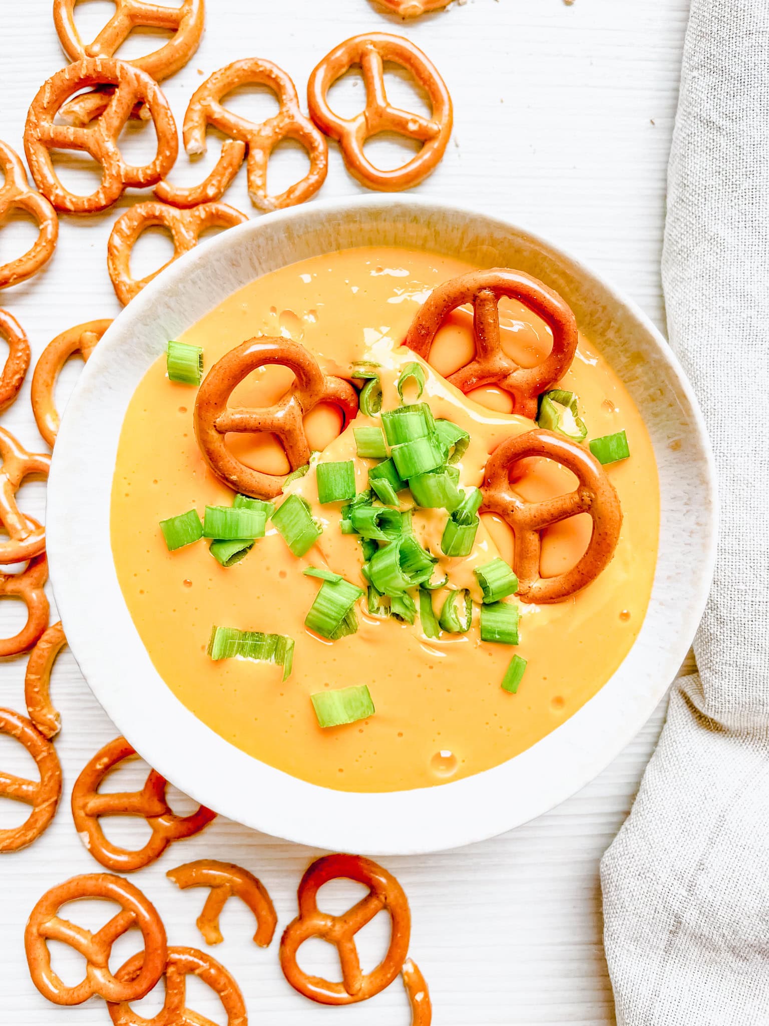 An overhead shot of the hot beer and cheese dip, topped with green onions and a few pretzels dipped into the bowl. More pretzels are scattered around the plate, creating a casual and inviting presentation.