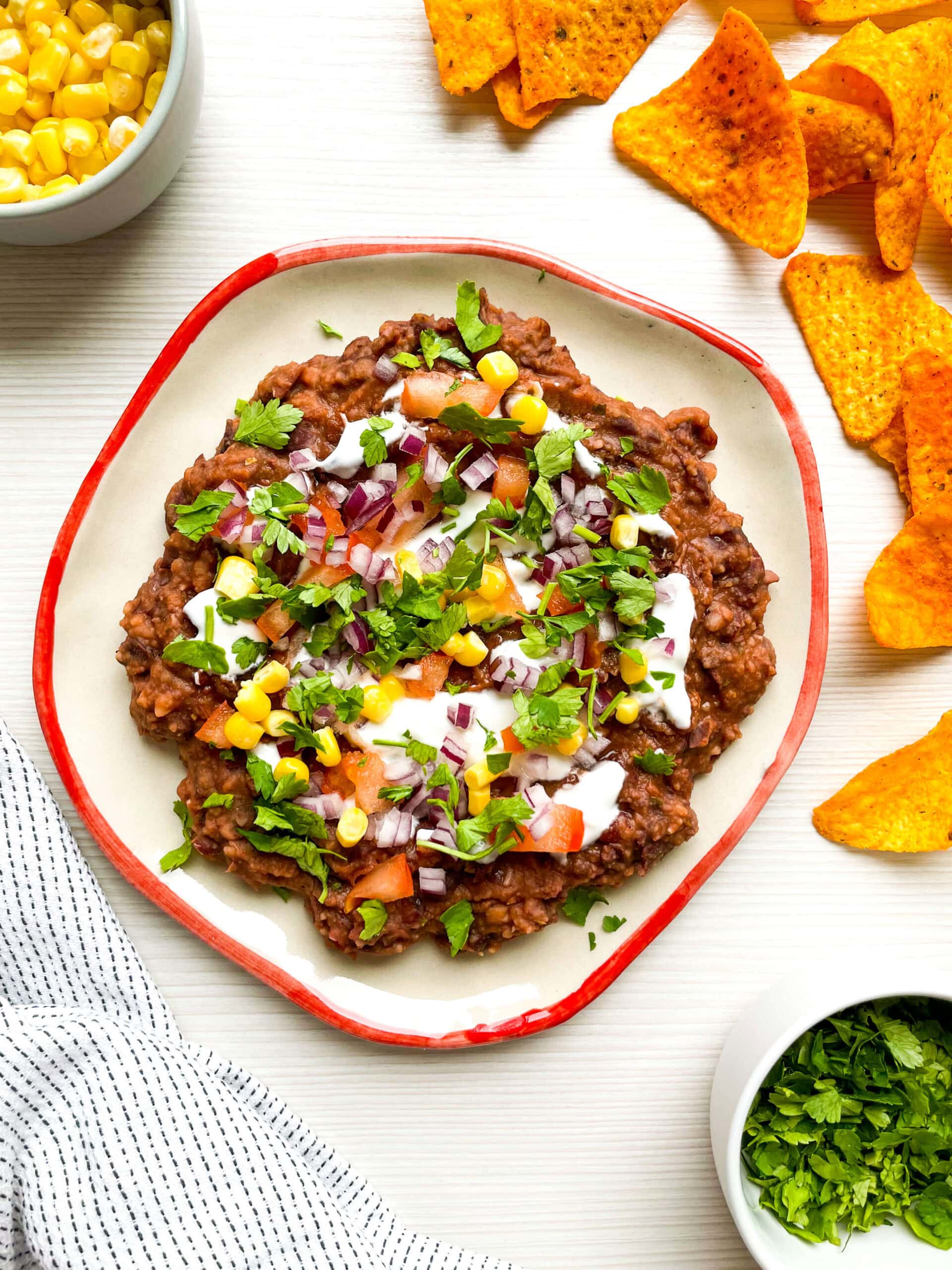 A top-down view of loaded black bean dip served on a ceramic plate with a red rim. The dish is surrounded by tortilla chips, a bowl of fresh corn, and a bowl of chopped cilantro on a white table.