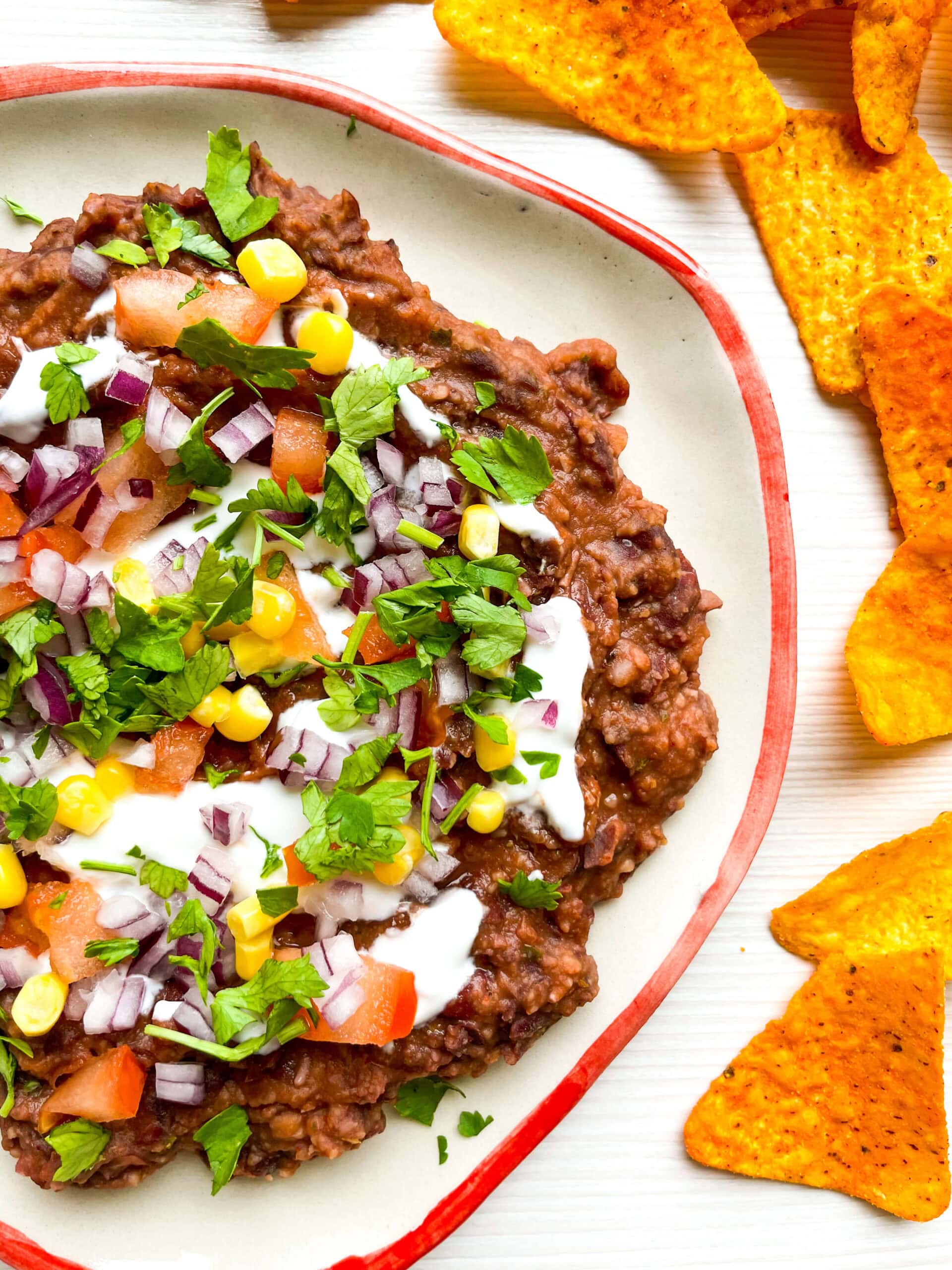 A slightly angled view of loaded black bean dip highlighting the colorful toppings, creamy drizzle of sour cream, and the orange tortilla chips arranged on the side.