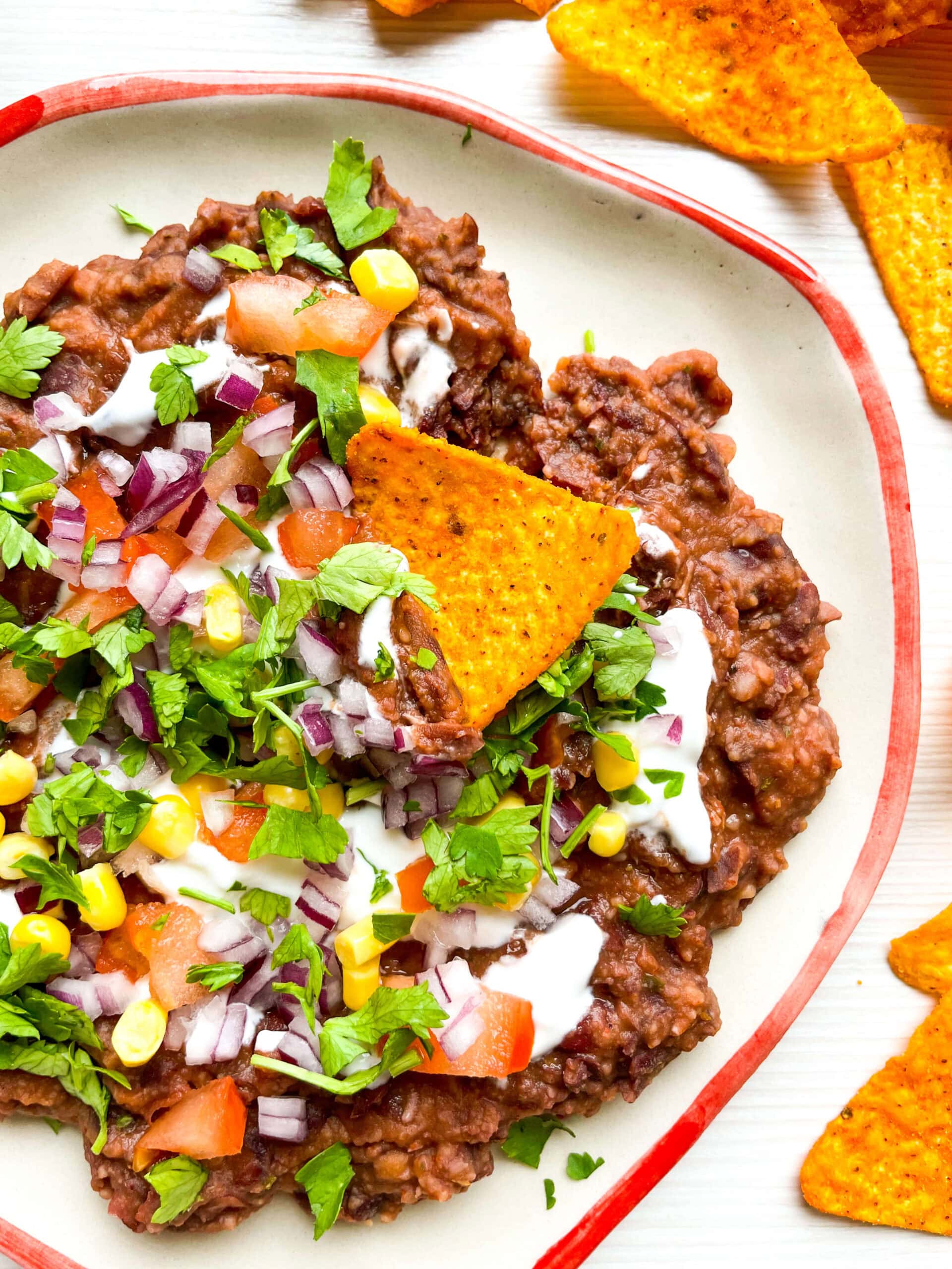 A plate of loaded black bean dip topped with fresh diced tomatoes, corn kernels, red onion, cilantro, and drizzled with sour cream. Bright orange tortilla chips are arranged around the plate for dipping.