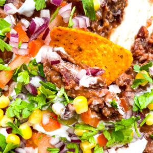 A close-up of loaded black bean dip showcasing the creamy texture of the beans, vibrant colors of diced tomatoes, corn, red onion, cilantro, and a crispy orange tortilla chip dipped into the dish.