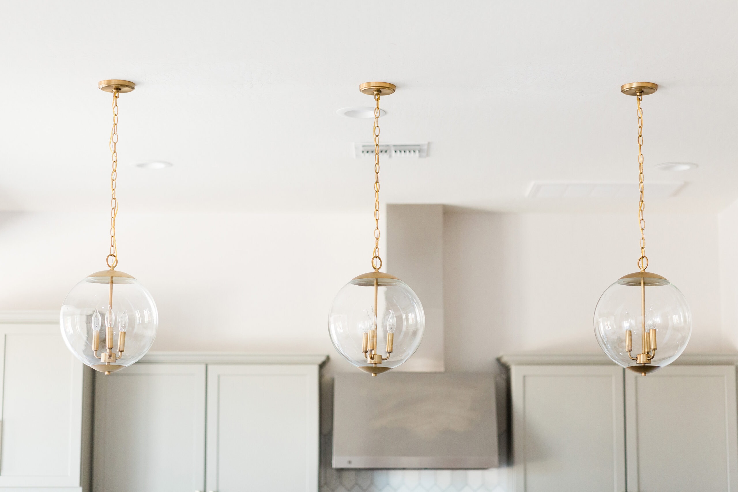 Three modern glass globe pendant lights with gold accents hanging from a white ceiling, creating a bright and elegant ambiance in a neutral-toned kitchen.
