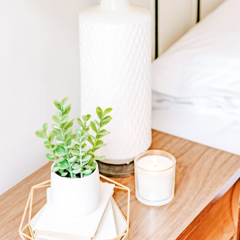 A cozy bedside table styled with a potted green plant, stacked books, a lit candle, and a white textured lamp, adding warmth and elegance to the bedroom.