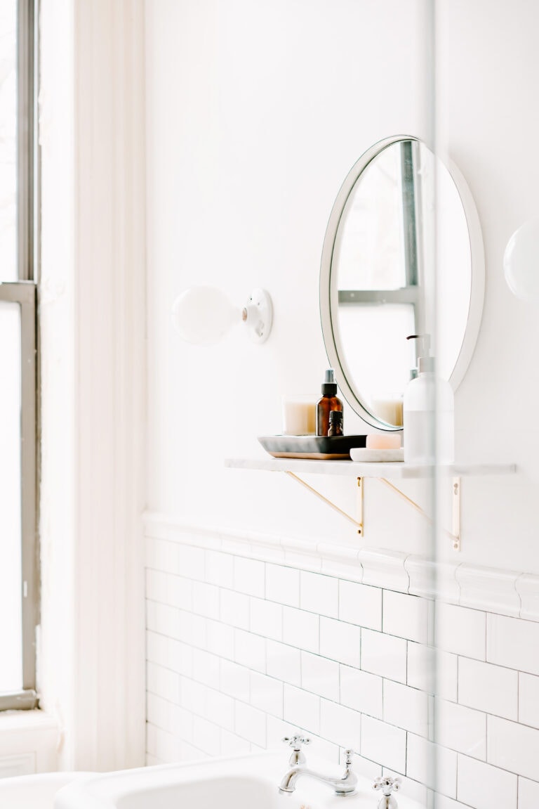A bright bathroom featuring a round mirror, a marble shelf with gold brackets, and neatly arranged essentials such as a brown bottle, a soap dish, and a candle. The white subway tiles and natural light create a clean, serene ambiance.
