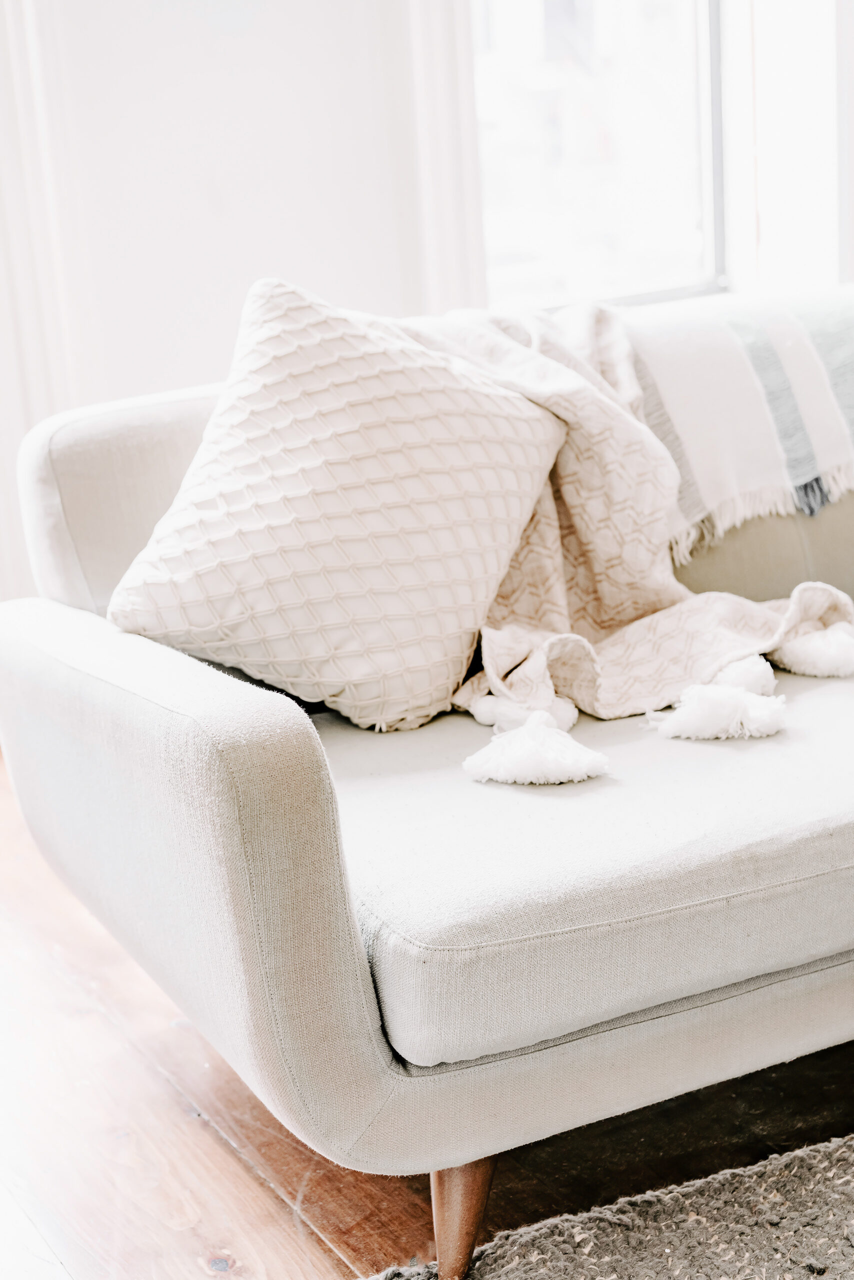 A cozy beige sofa styled with a textured throw blanket and a quilted accent pillow, creating a warm and inviting living room vibe. Soft natural light streams through the window in the background.