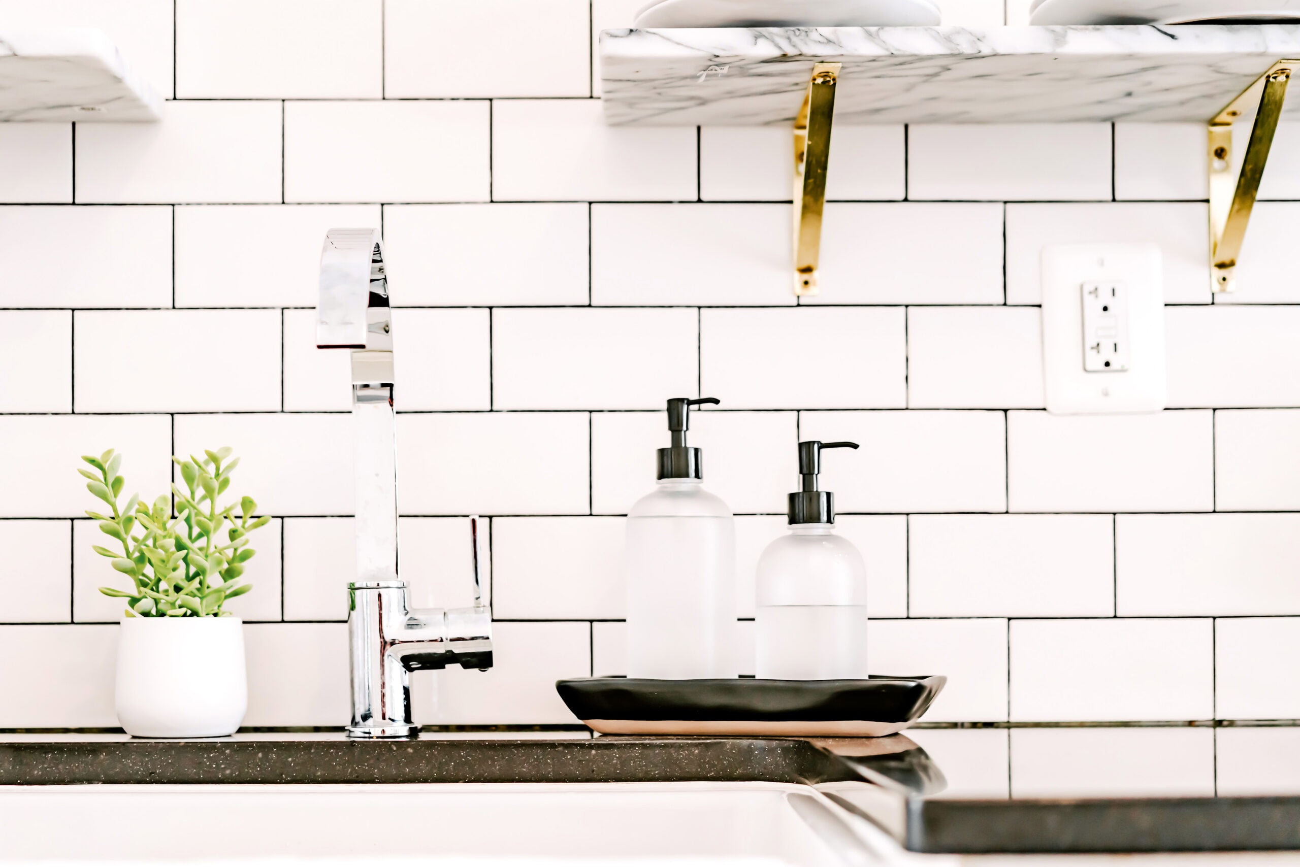 A modern kitchen sink area with a sleek chrome faucet, white subway tile backsplash, and minimalistic decor, including a small green potted plant and two frosted soap dispensers on a black tray.