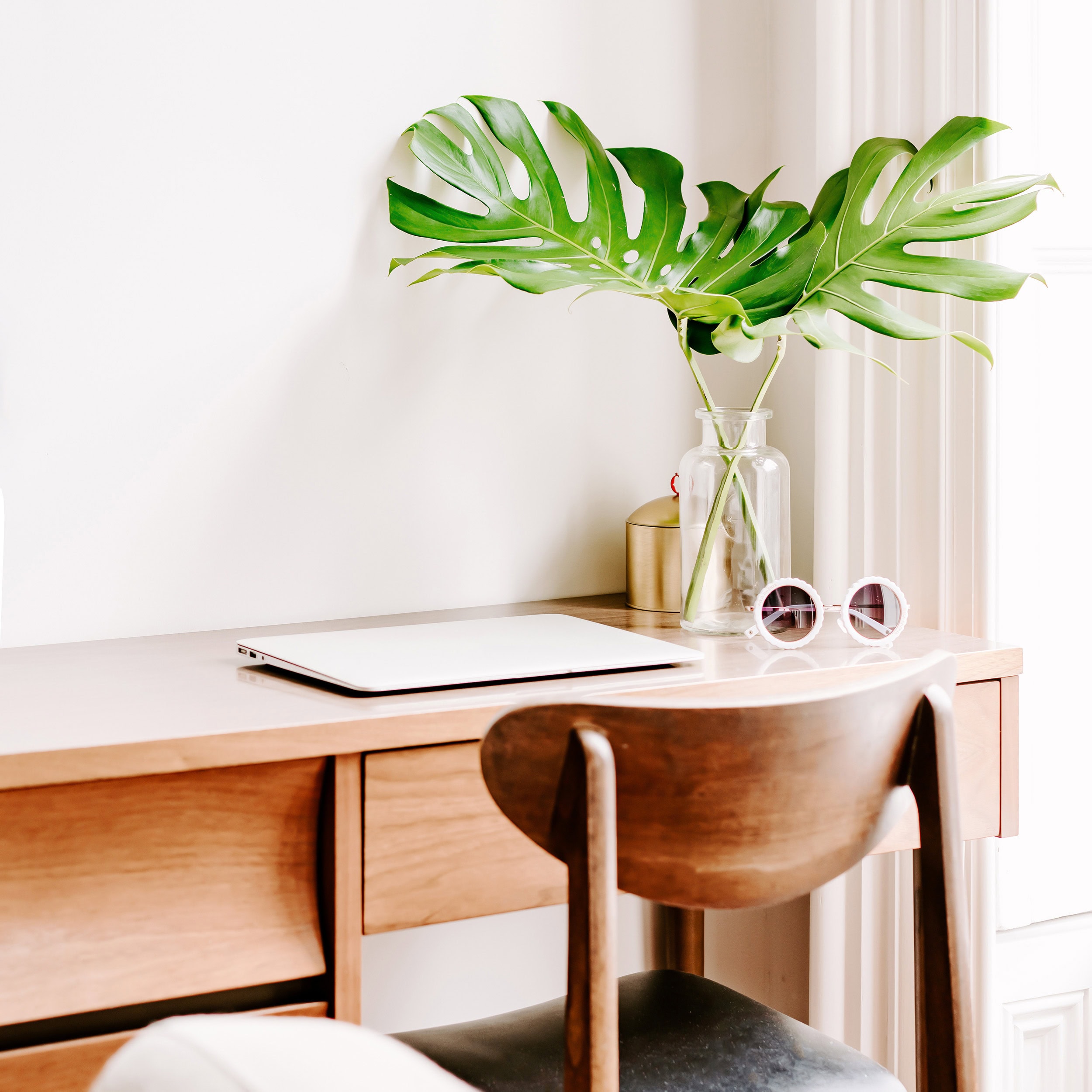 A minimalistic wooden desk with a sleek laptop, a glass vase holding tropical monstera leaves, and chic sunglasses, creating a fresh and stylish workspace.