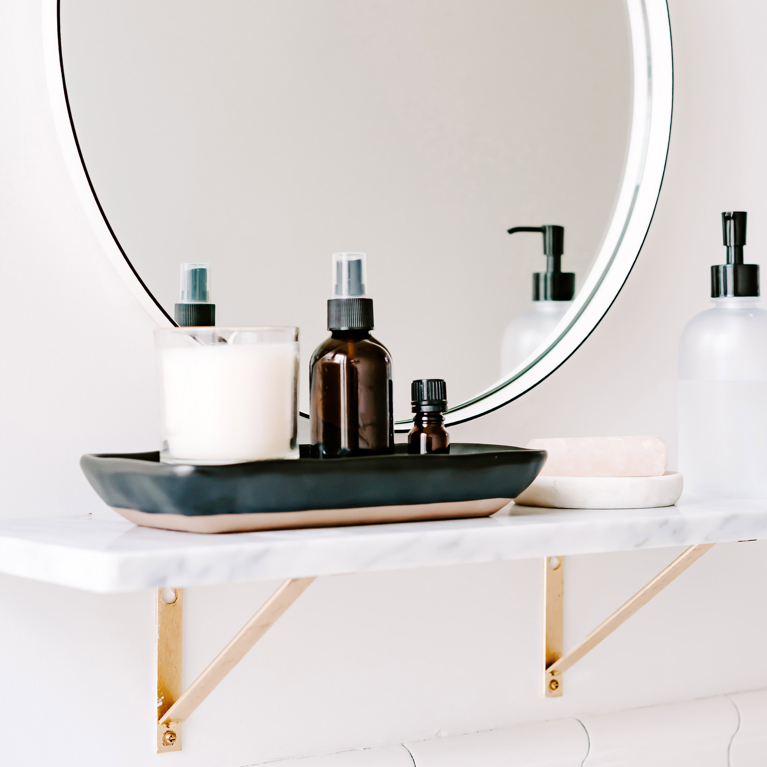 A beautifully organized bathroom shelf with a round mirror and a marble tray holding candles, amber glass bottles, and soap, styled to feel serene and spa-like