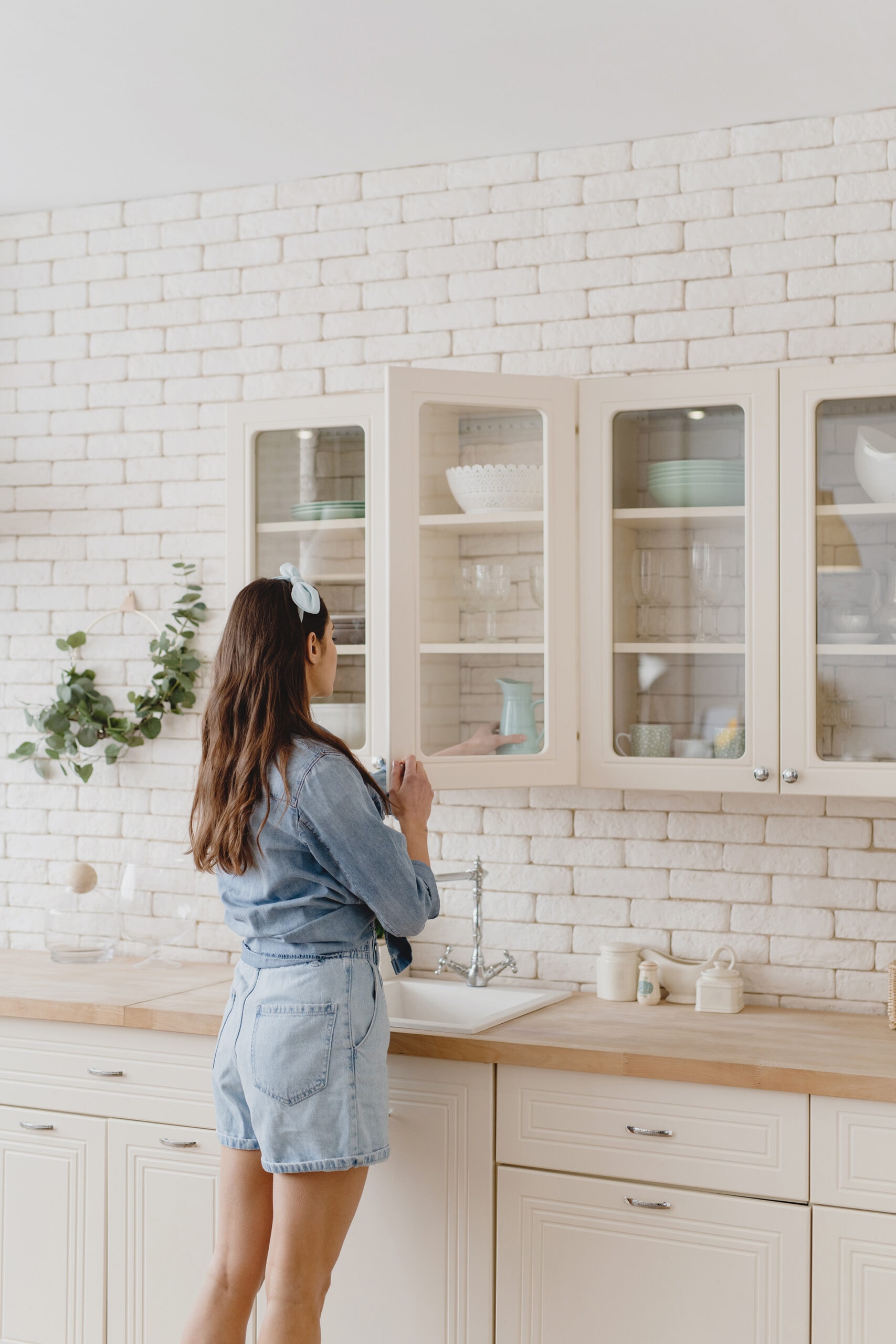A woman in a denim outfit reaching into a glass-front kitchen cabinet to organize dishes. The bright kitchen has a white brick backsplash and light wooden countertops, creating a cozy and tidy atmosphere.