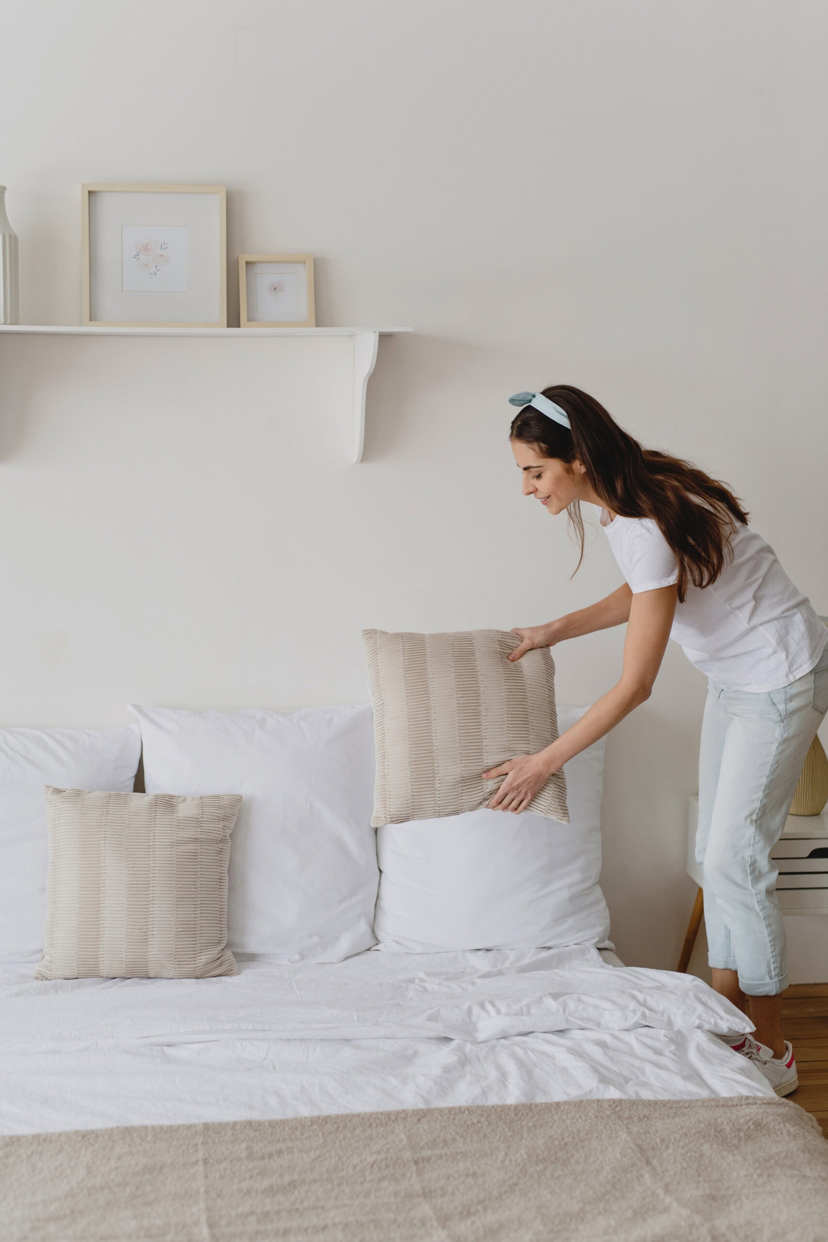 A woman in casual white and light denim attire arranging neutral throw pillows on a neatly made bed. The room has a calming aesthetic with white walls, simple decor, and framed prints on a shelf above the bed.
