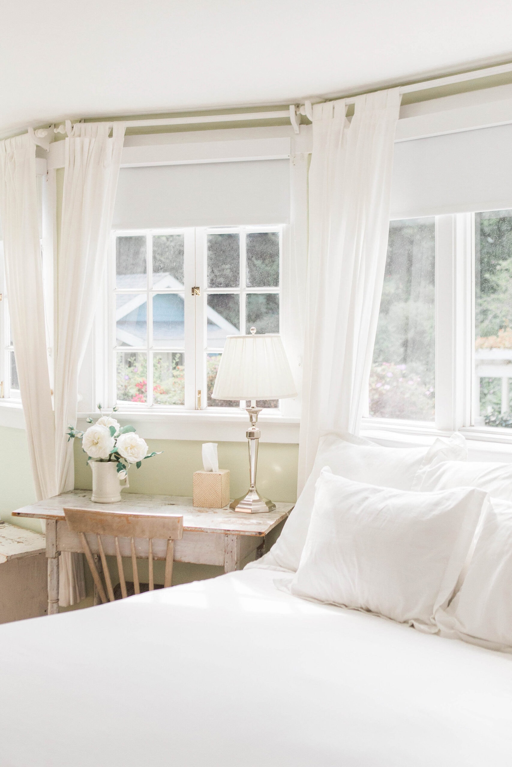 A charming corner of a bedroom with a white bed beside a rustic wooden desk. The desk features a silver lamp, a vase of white flowers, and a tissue box, illuminated by natural light streaming through the surrounding windows.