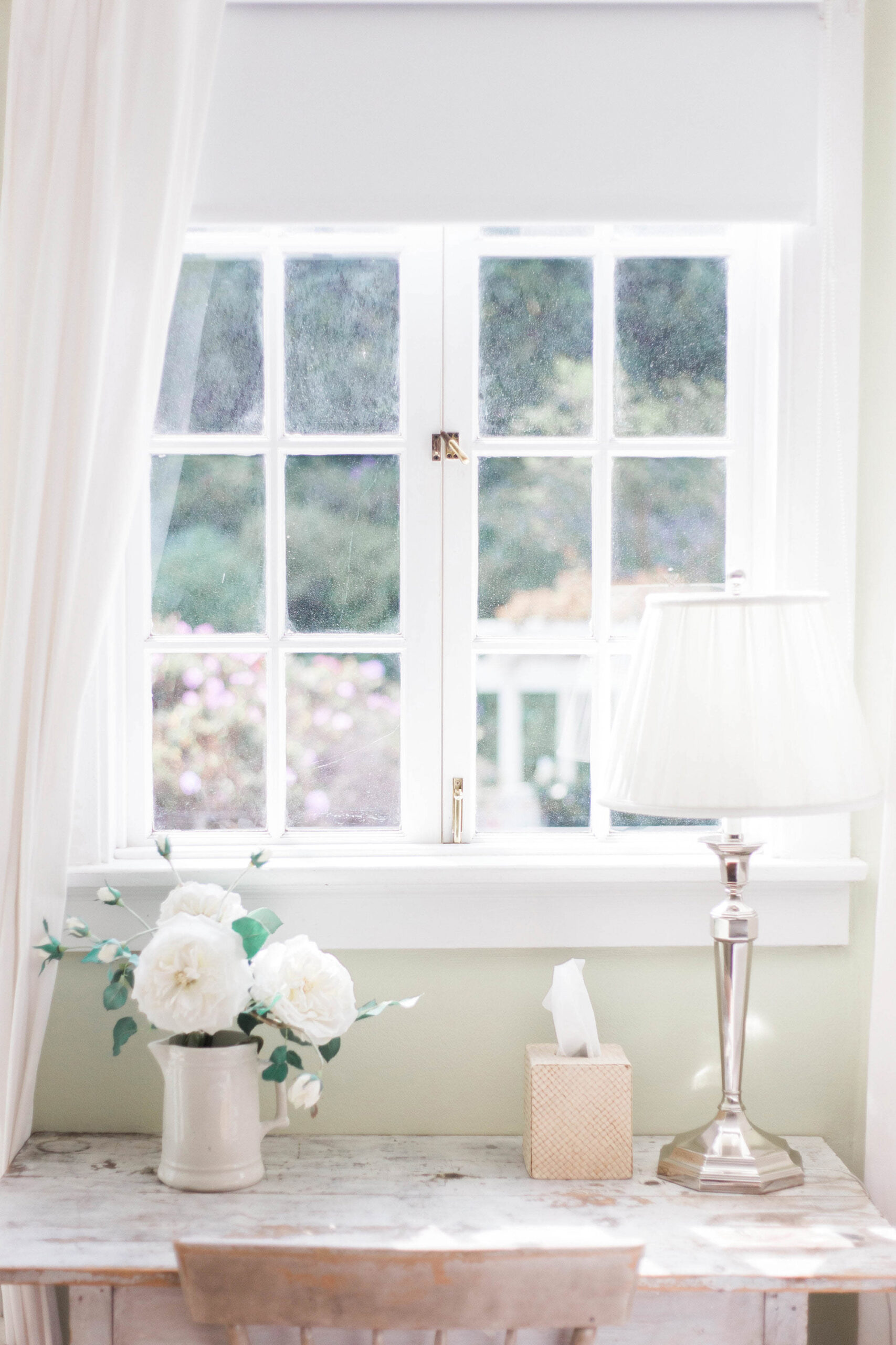 A rustic desk by a window with a white ceramic vase of white roses and eucalyptus, a woven tissue box, and a polished silver lamp. The soft green walls and white curtains create a tranquil ambiance.