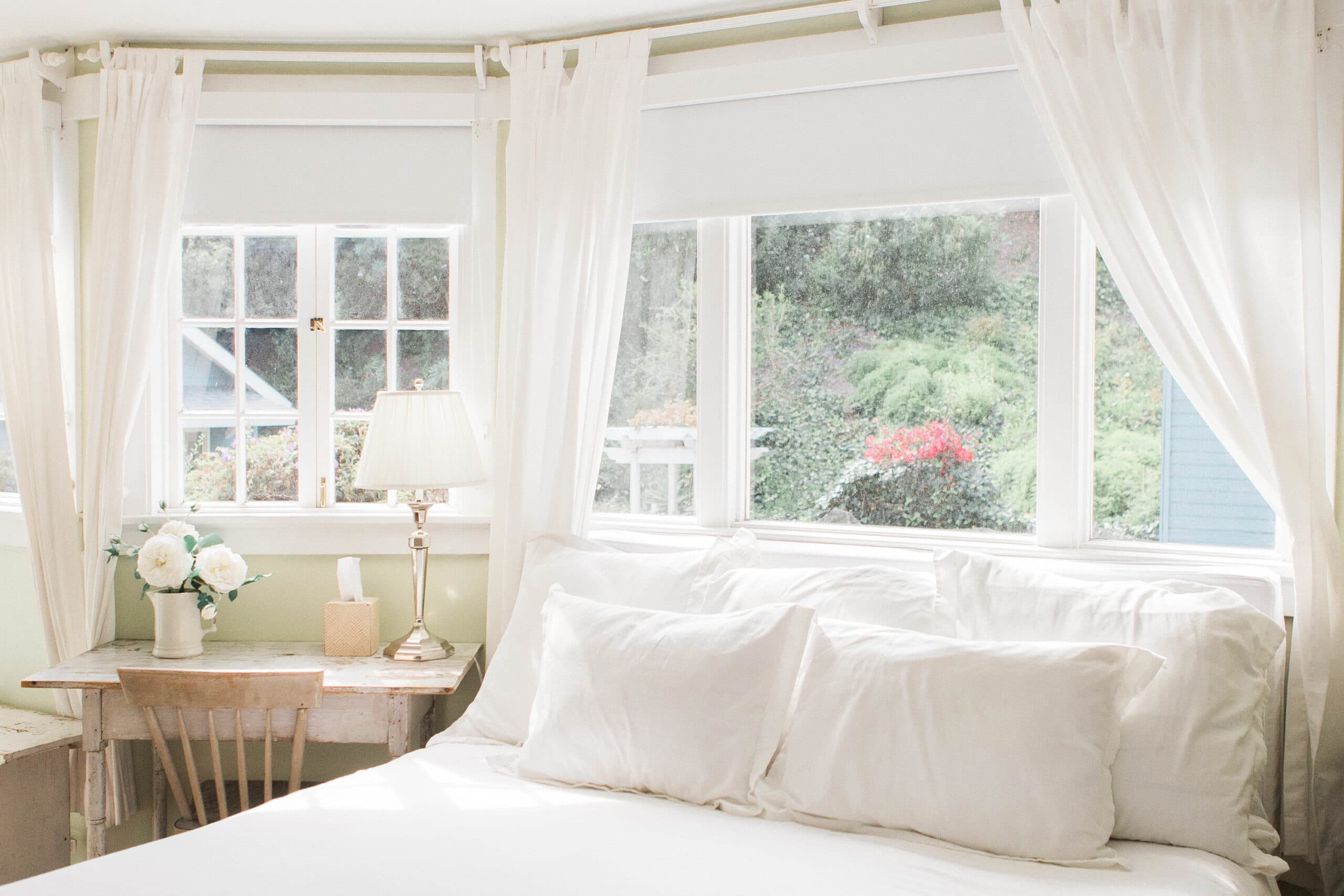 A bright bedroom scene showcasing a bed with white linens and multiple pillows near a set of windows draped with airy white curtains. Outside, lush greenery and vibrant flowers peek through.