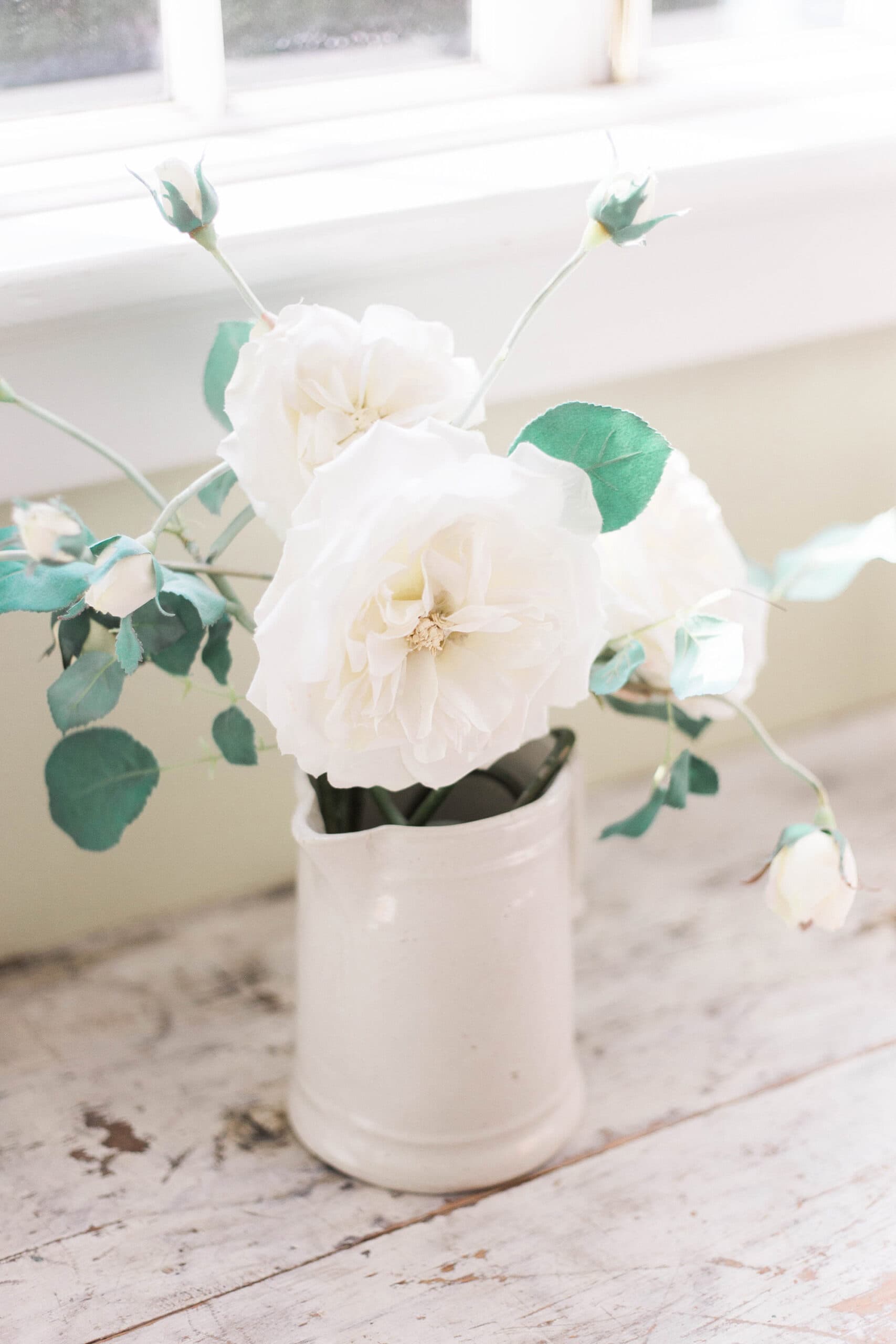 A close-up of a white ceramic vase holding white roses and eucalyptus on a distressed wooden desk. The soft natural light highlights the delicate petals and vintage aesthetic.