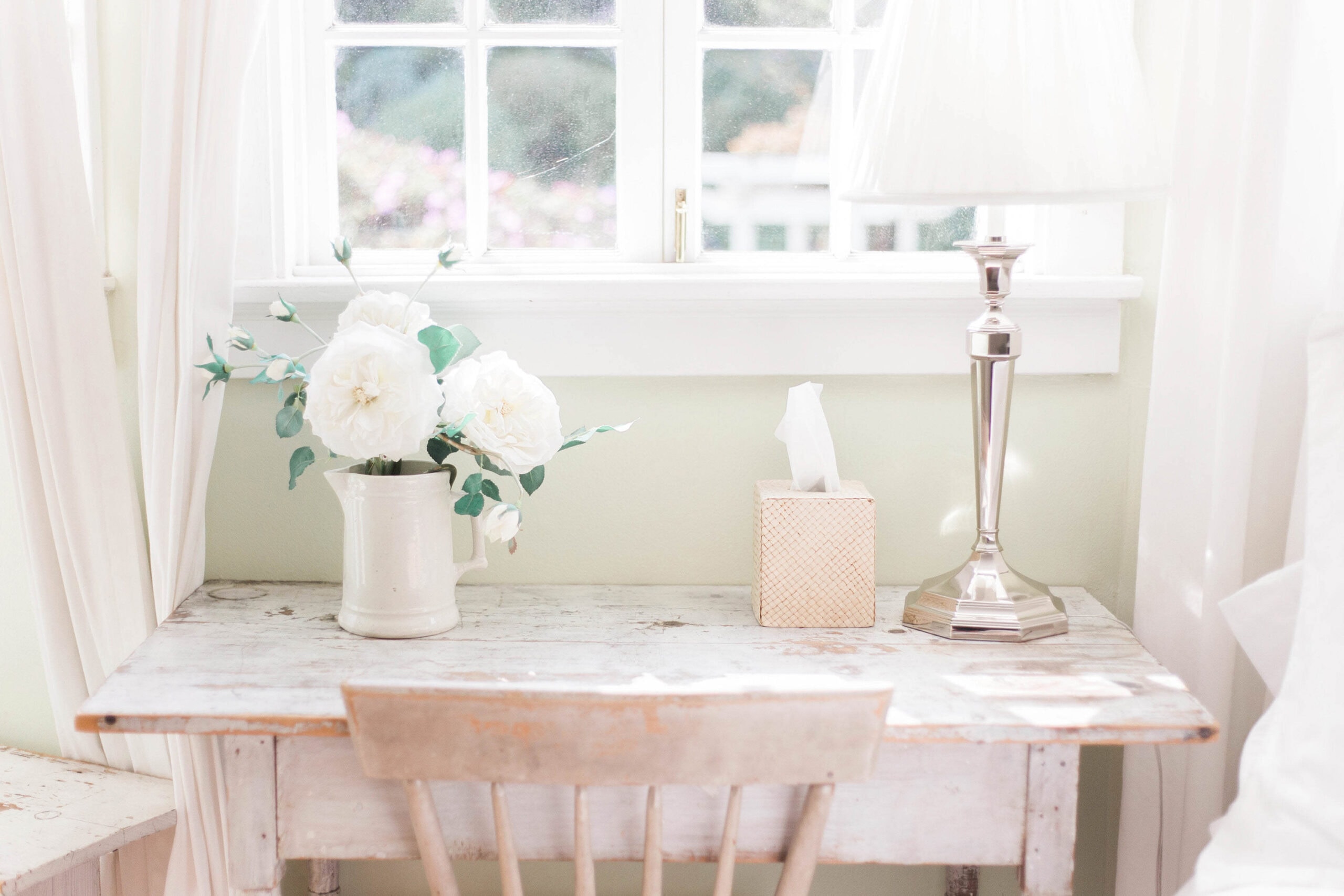 A cozy desk setup featuring a white ceramic vase with white roses and eucalyptus, a tissue box, and a silver table lamp. The desk is positioned in front of a window with soft natural light filtering through white curtains.