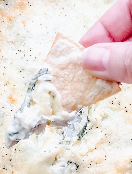 A hand dipping a crispy tortilla chip into the warm and creamy spinach and artichoke dip, highlighting the gooey cheese and artichoke chunks