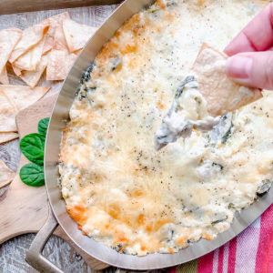 A silver dish filled with bubbly, cheesy spinach and artichoke dip with a chip being dipped in, surrounded by homemade tortilla chips and fresh spinach leaves on a rustic table.