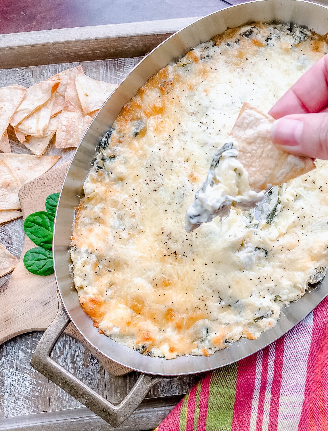 A silver dish filled with bubbly, cheesy spinach and artichoke dip with a chip being dipped in, surrounded by homemade tortilla chips and fresh spinach leaves on a rustic table.