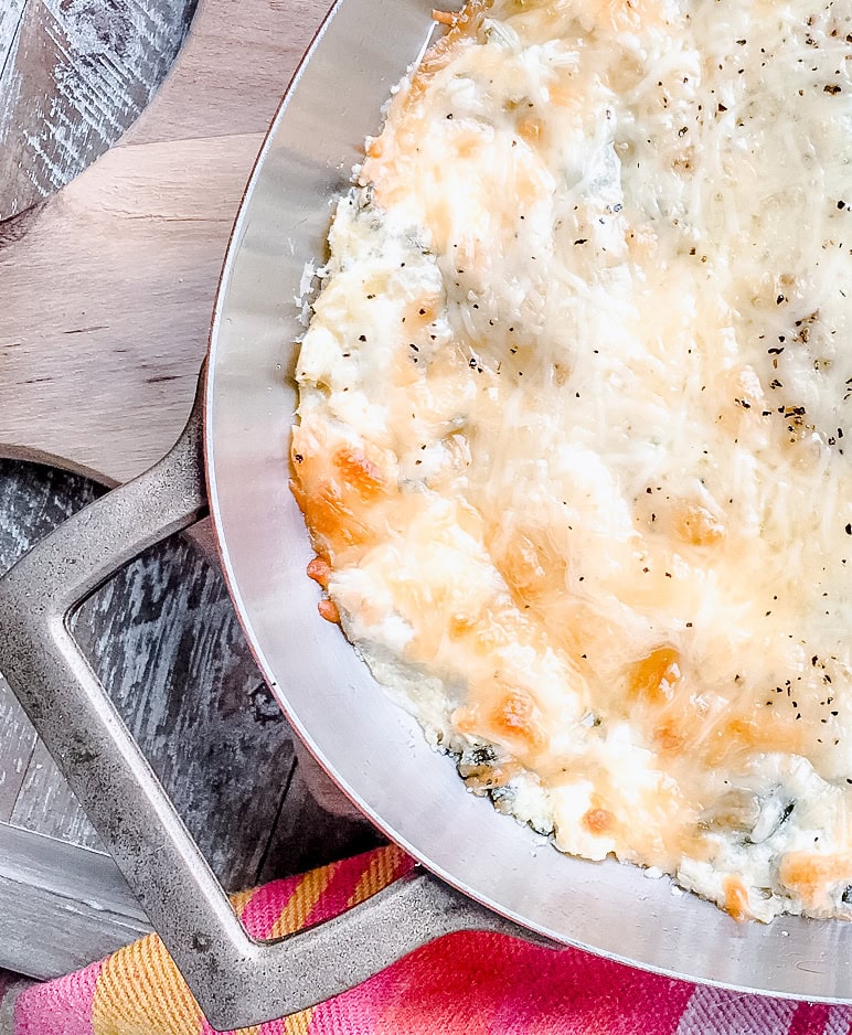 A close-up view of golden-brown, cheesy spinach and artichoke dip baked to perfection in a silver dish, set on a rustic wooden table with a vibrant striped napkin.