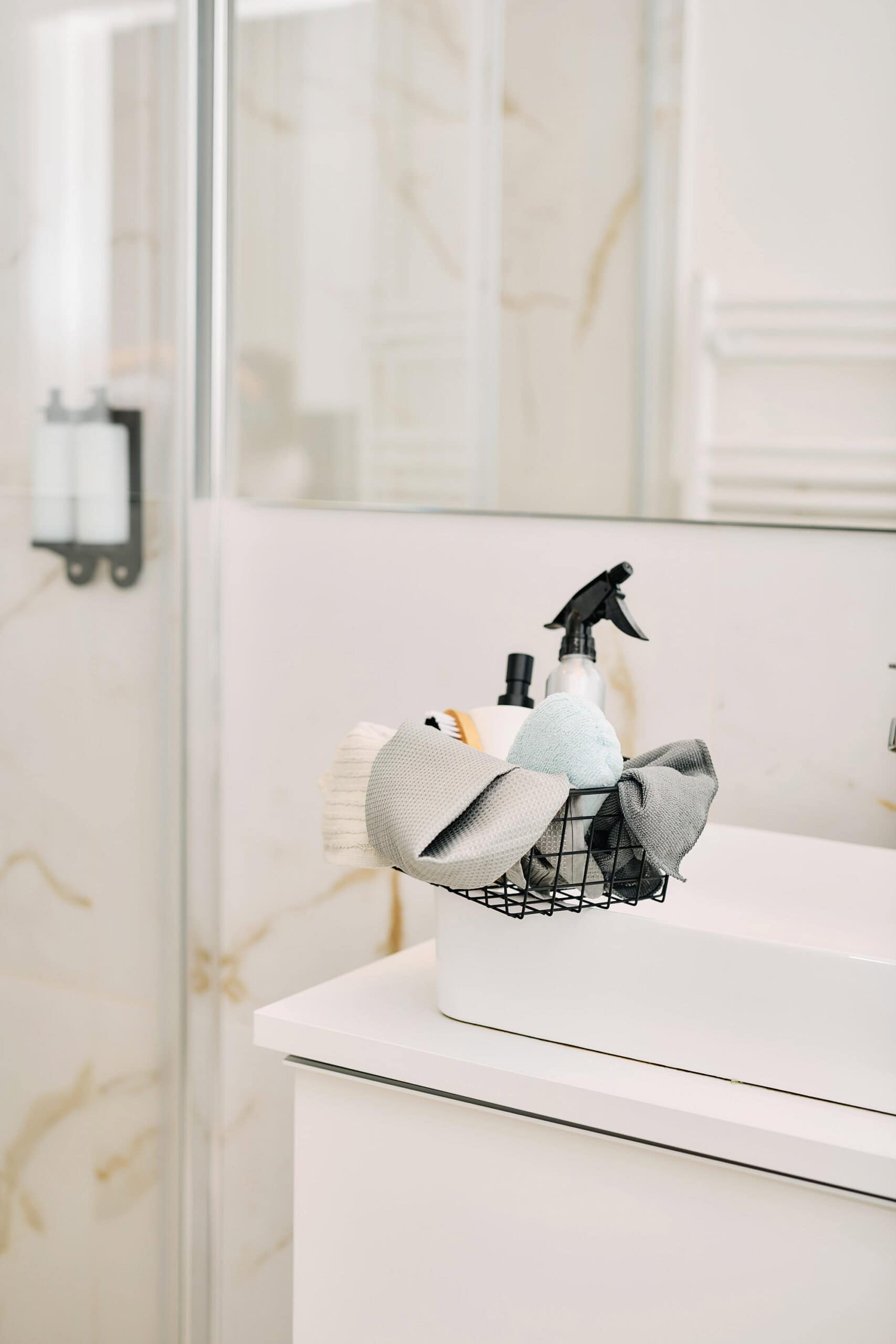 A minimalist bathroom scene with a white sink, a marble backsplash, and a black wire basket holding cleaning tools like a spray bottle, towels, and a sponge.