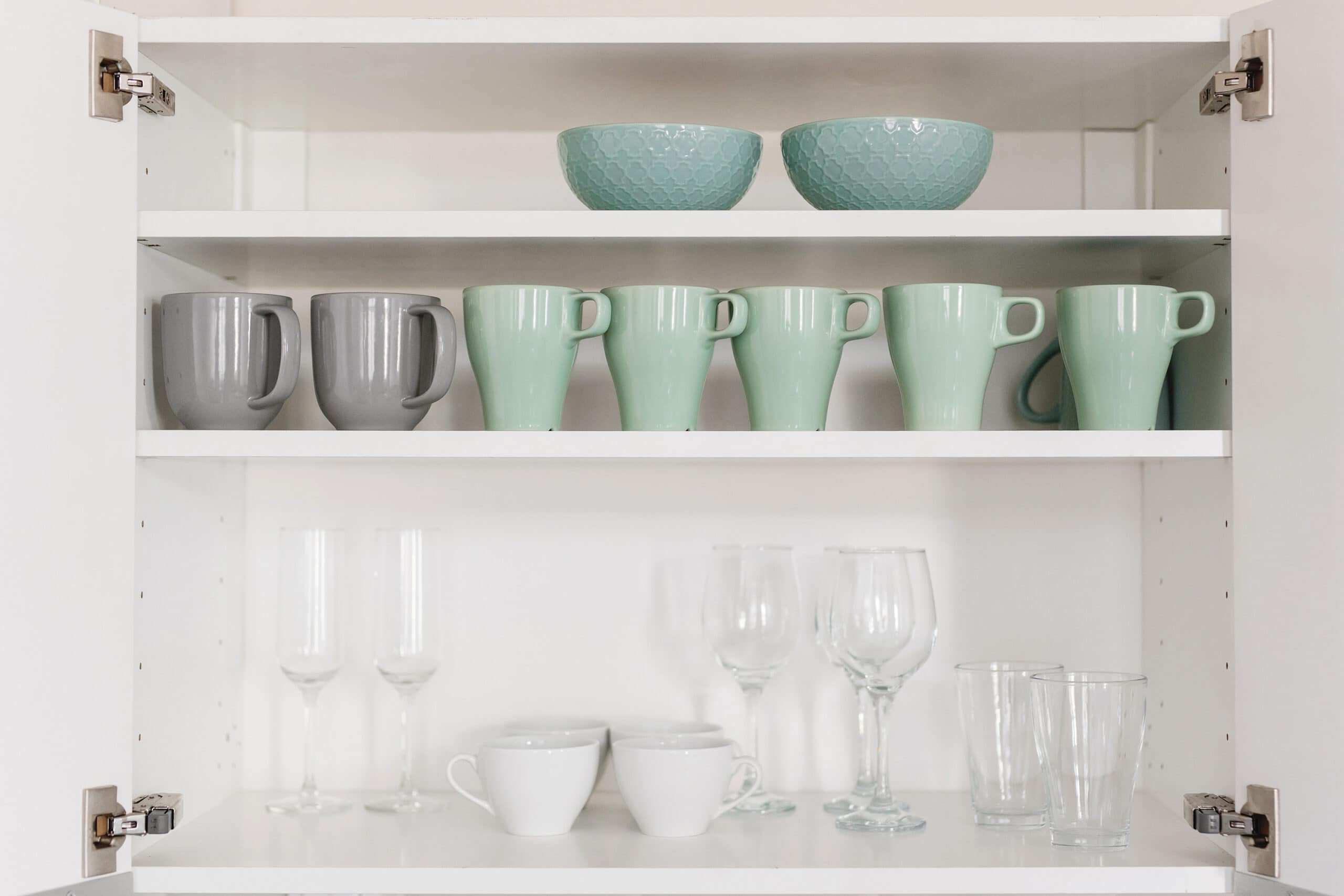 An open kitchen cabinet with neatly arranged dishware, featuring mint green and gray mugs, textured green bowls, white teacups, and a selection of wine and champagne glasses, creating an organized and aesthetic look.