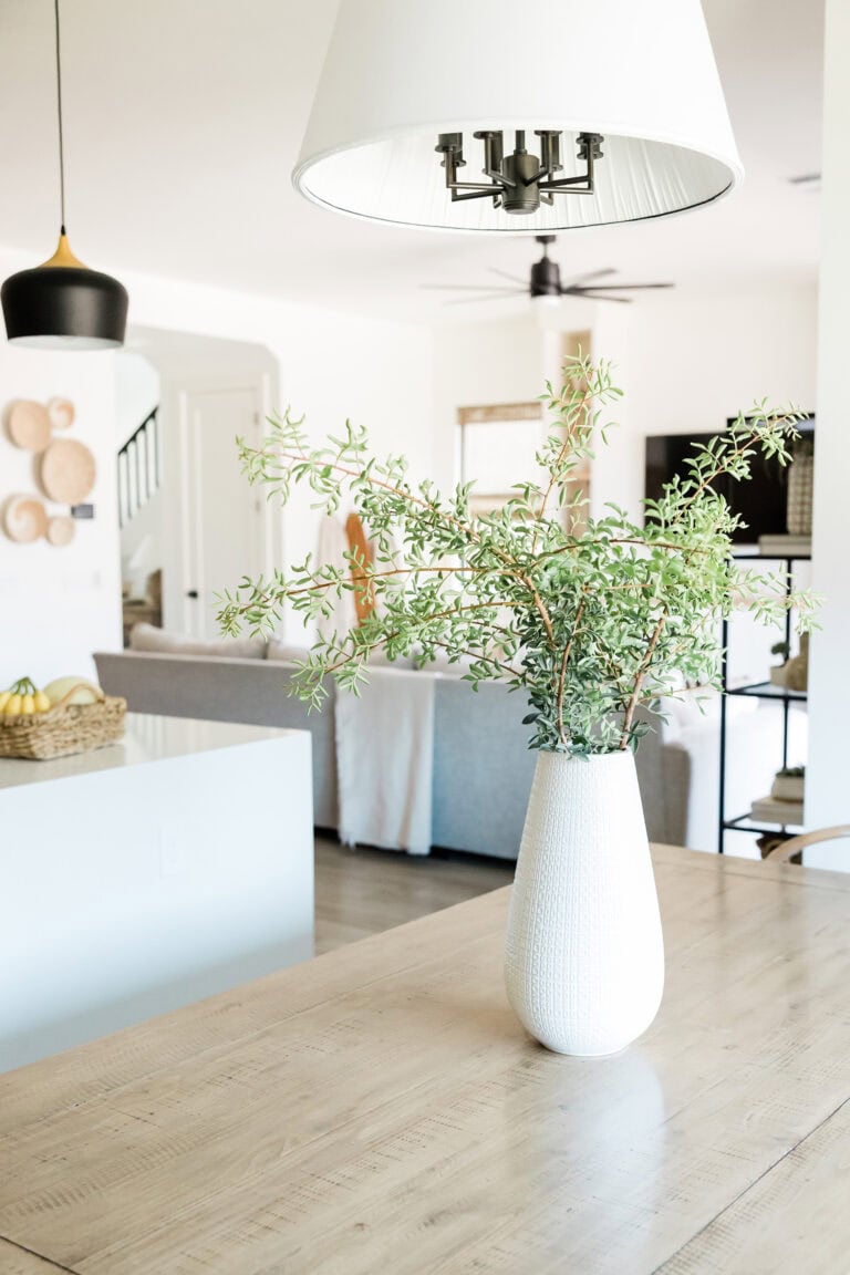 A white vase with fresh greenery displayed on a natural wood dining table, surrounded by a light and airy open-concept living spa