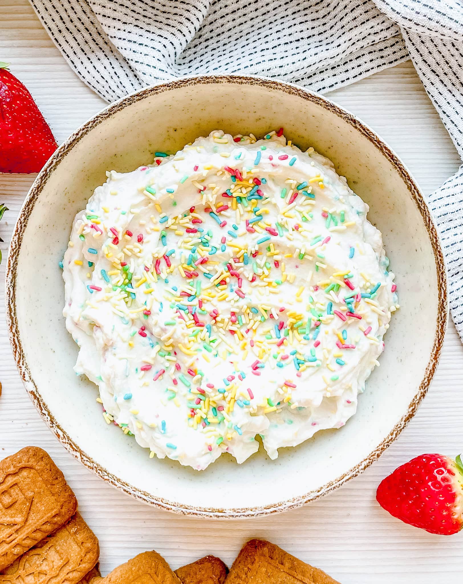 A top-down view of a bowl filled with a creamy dip, sprinkled with bright pastel-colored rainbow sprinkles, surrounded by speculoos cookies and fresh strawberries on a clean white background.