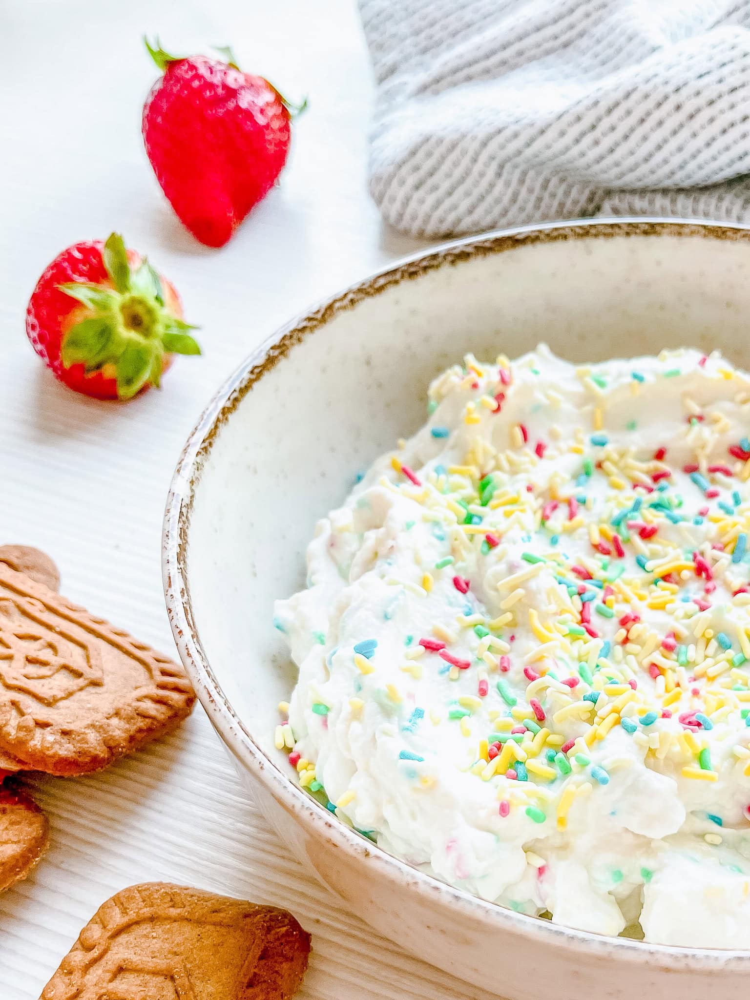 A ceramic bowl filled with a creamy white dip, topped with colorful rainbow sprinkles. The bowl is placed on a white surface next to fresh strawberries and rectangular speculoos cookies.