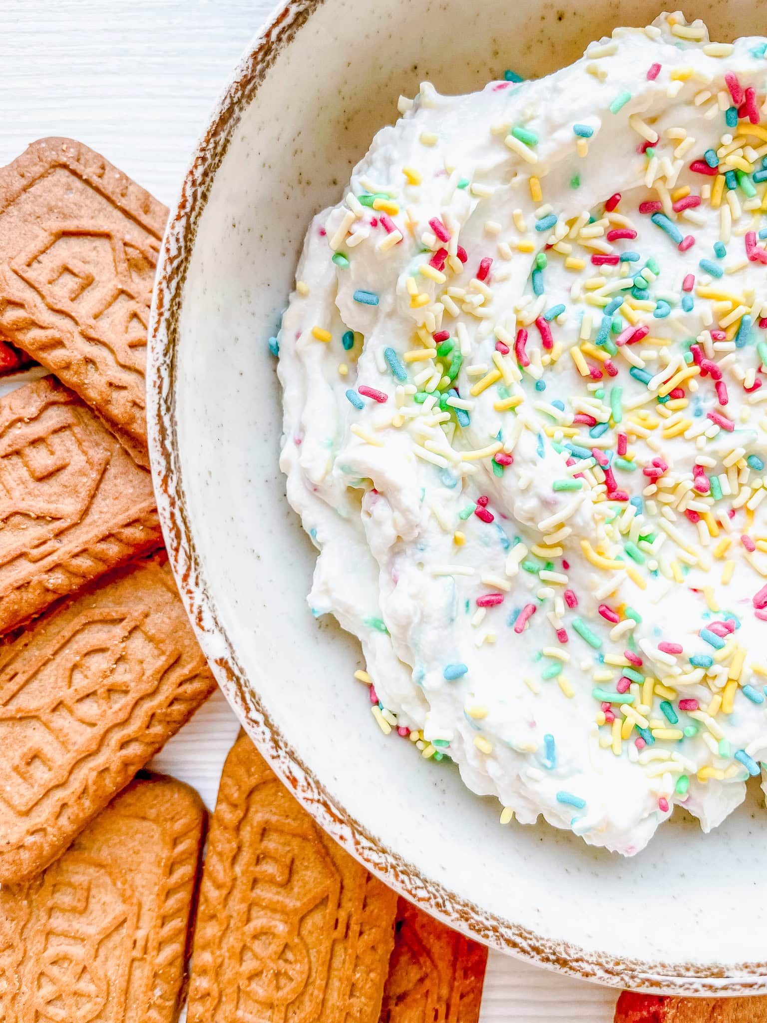 A close-up shot of a ceramic bowl containing a fluffy white dip, decorated with pastel-colored rainbow sprinkles, surrounded by speculoos cookies on a white surface.