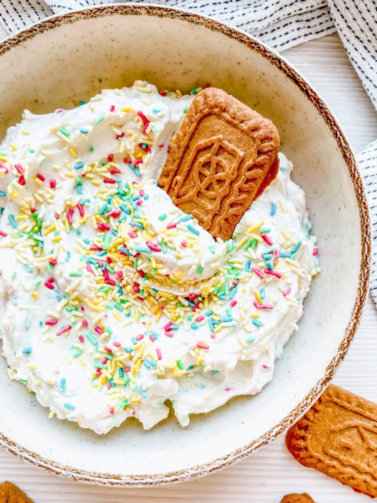 A ceramic bowl of white whipped dip with rainbow sprinkles, with a speculoos cookie inserted into the dip for a playful presentation. The background features a light cloth and additional cookies.