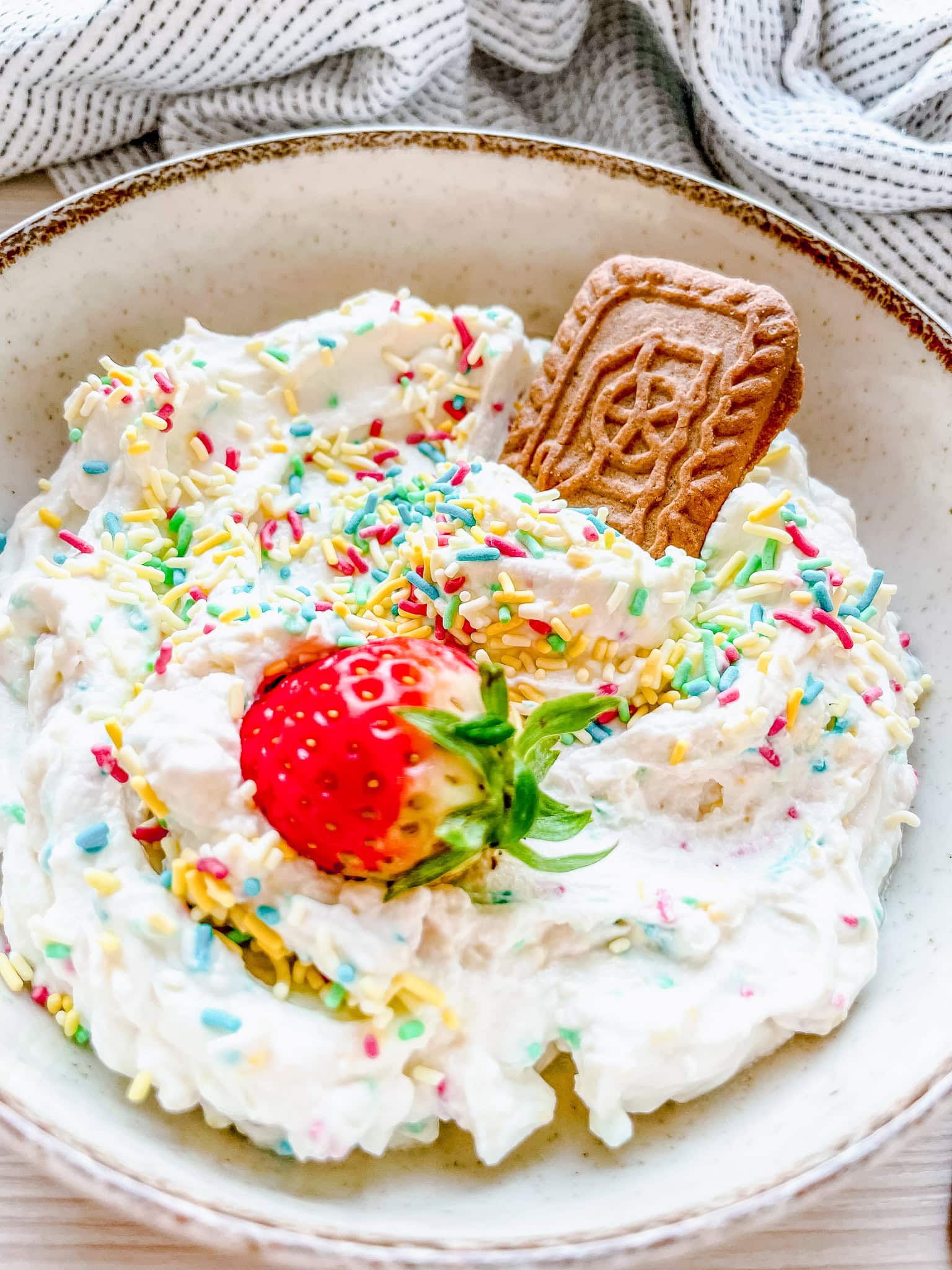 A festive bowl of whipped dip, sprinkled with colorful sprinkles, accompanied by a fresh strawberry and a speculoos cookie partially dipped. The presentation is cozy and inviting.