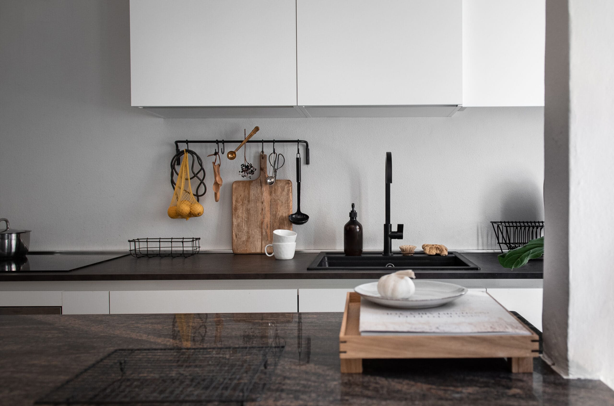 A sleek and modern kitchen counter with dark surfaces, a wooden cutting board, garlic, and utensils hanging from a black rail. A minimalistic aesthetic with functional design elements.