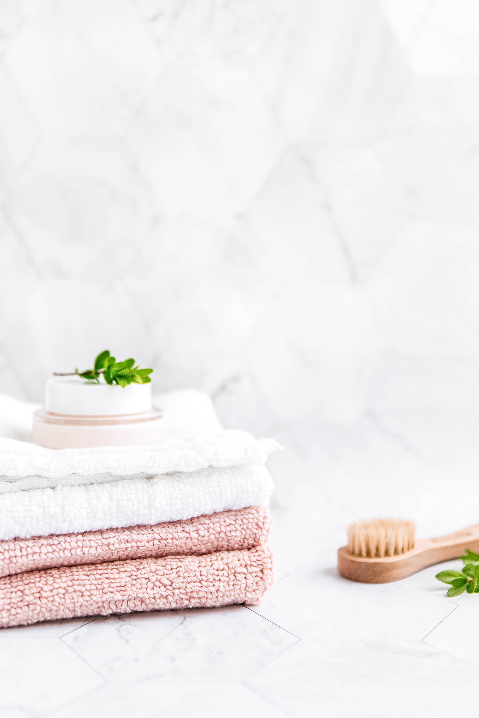 A folded stack of white and blush pink towels with a small jar of cream on top, set against a marble-tiled background. A wooden dry brush and a sprig of greenery complete the clean and cozy look.