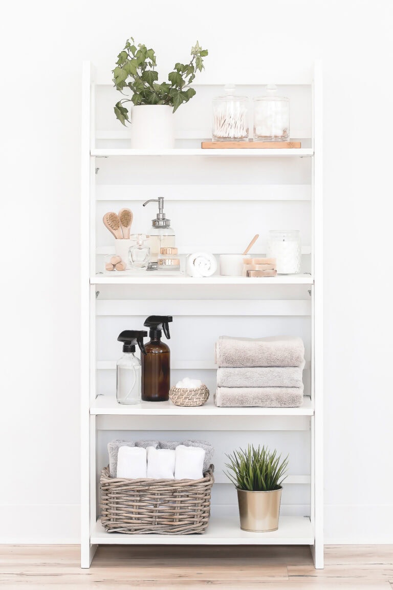 A white shelving unit styled with neatly arranged self-care and cleaning items, including potted greenery, jars of cotton swabs and cotton balls, folded towels, and amber cleaning spray bottles. A woven basket with rolled towels sits at the bottom, adding a touch of warmth.