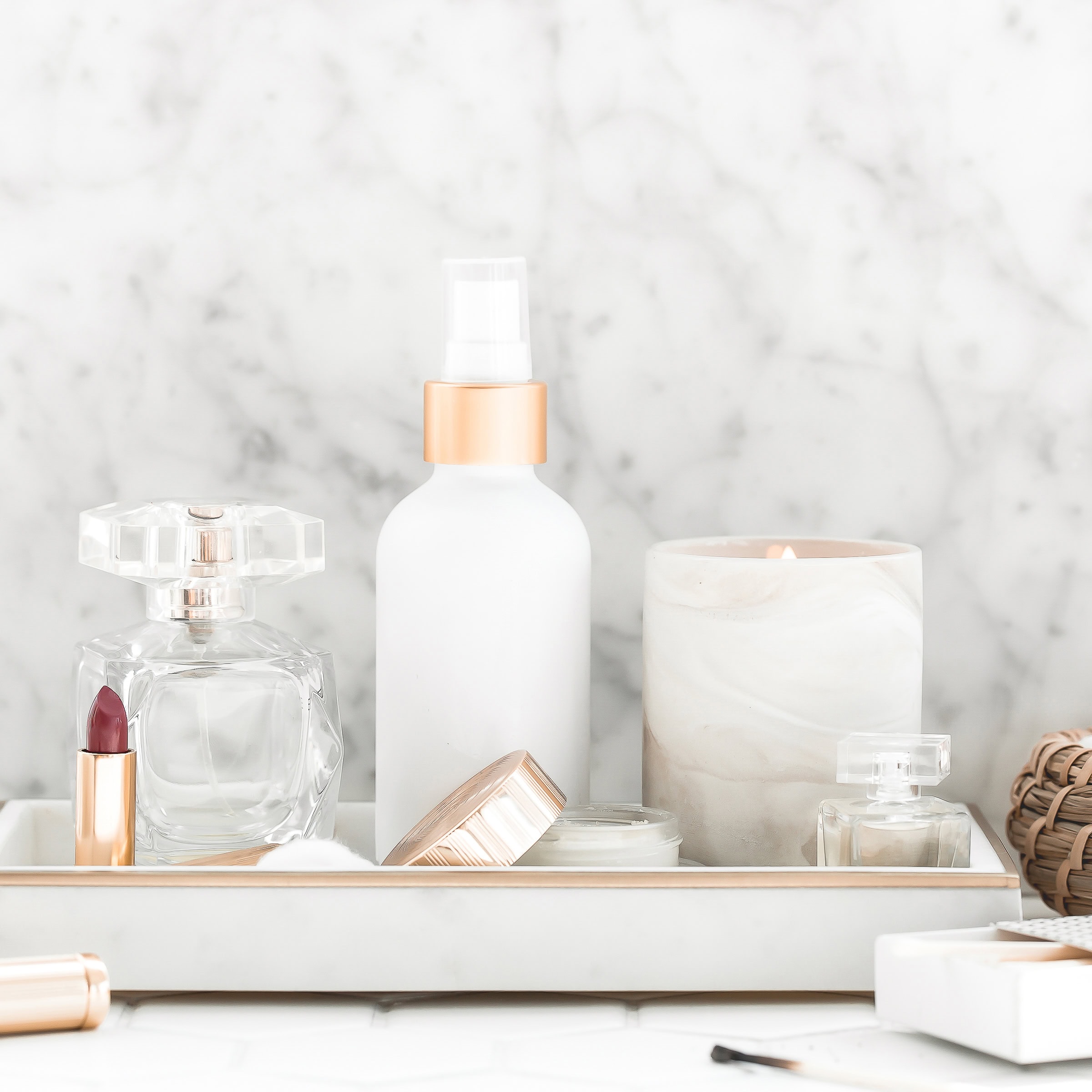 A marble tray holding elegant self-care essentials, including a crystal perfume bottle, a white spray bottle with a gold cap, a small candle in a marble holder, and an open lipstick. The background features a luxurious marble surface, creating a chic and serene vibe.