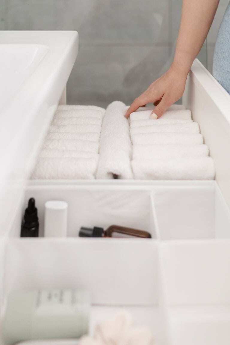 A person’s hand neatly arranging rolled white towels in an organized drawer. The drawer also features compartments containing bottles and other small items, suggesting a clean and minimalist bathroom setup.