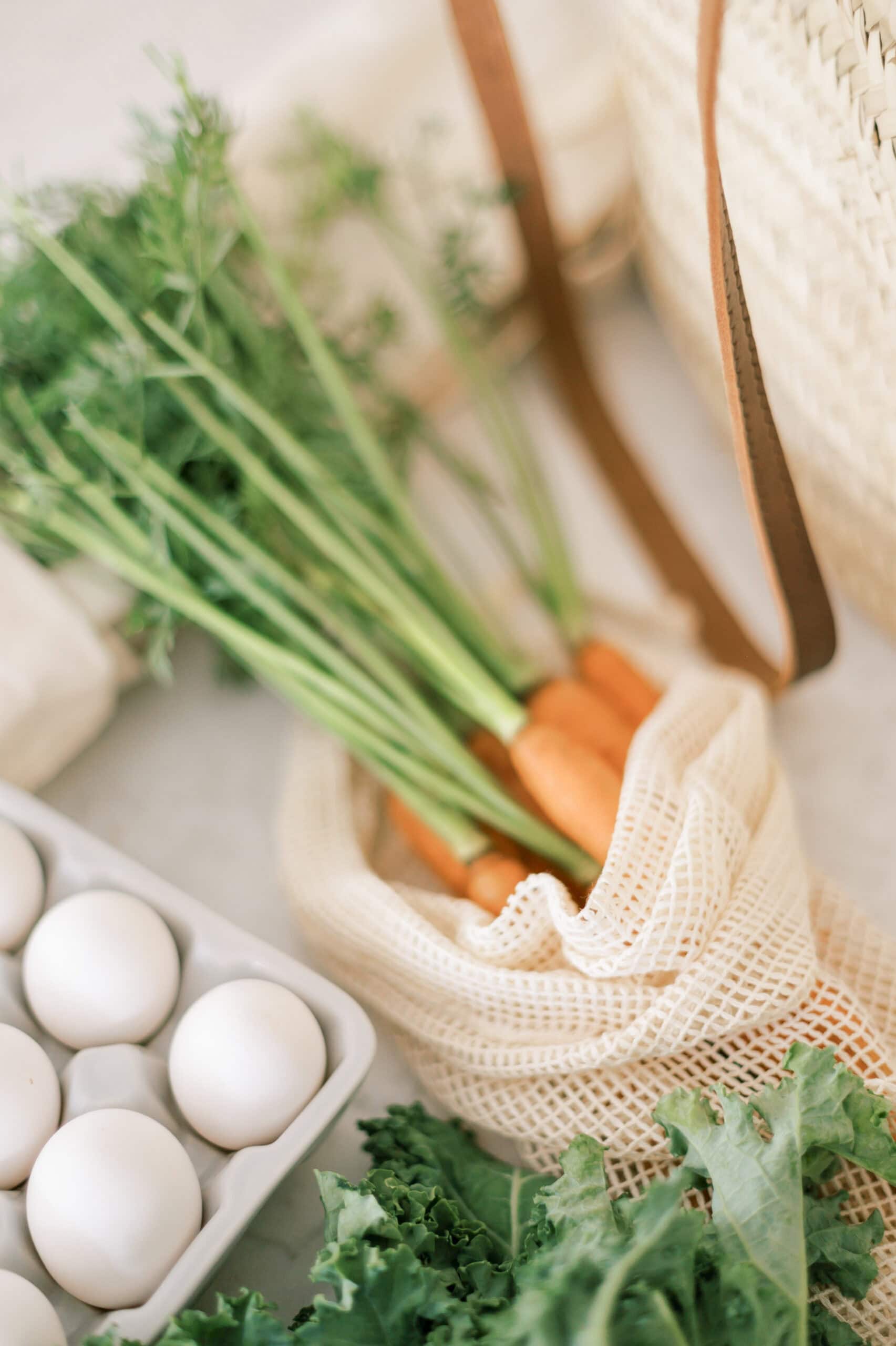 A close-up of a sustainable grocery setup featuring fresh carrots with greens in a reusable mesh bag, a carton of white eggs, and kale on a light countertop.