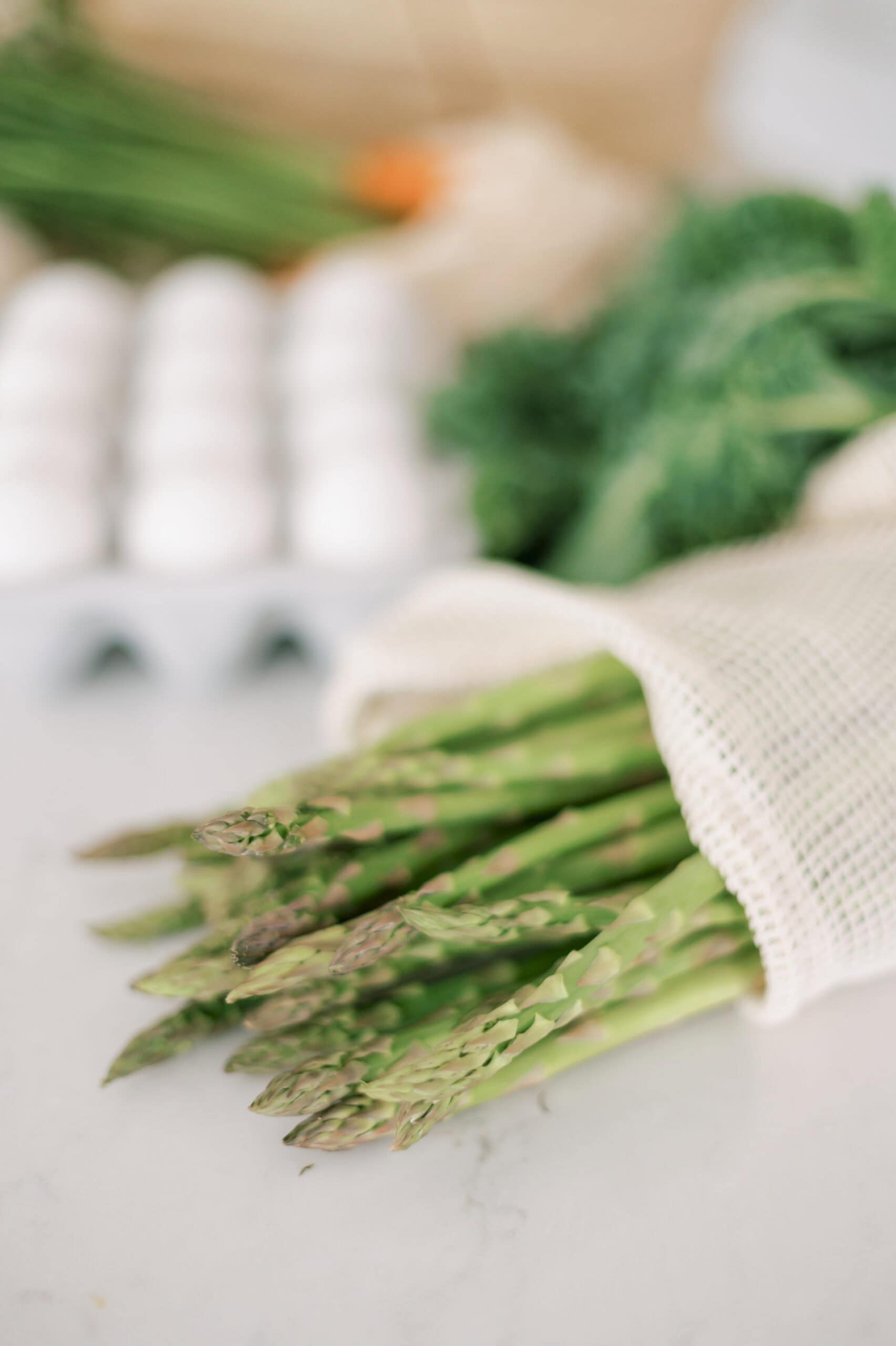 Fresh asparagus bundled in a reusable mesh bag, accompanied by a blurred background featuring a carton of white eggs and leafy greens on a light kitchen countertop.